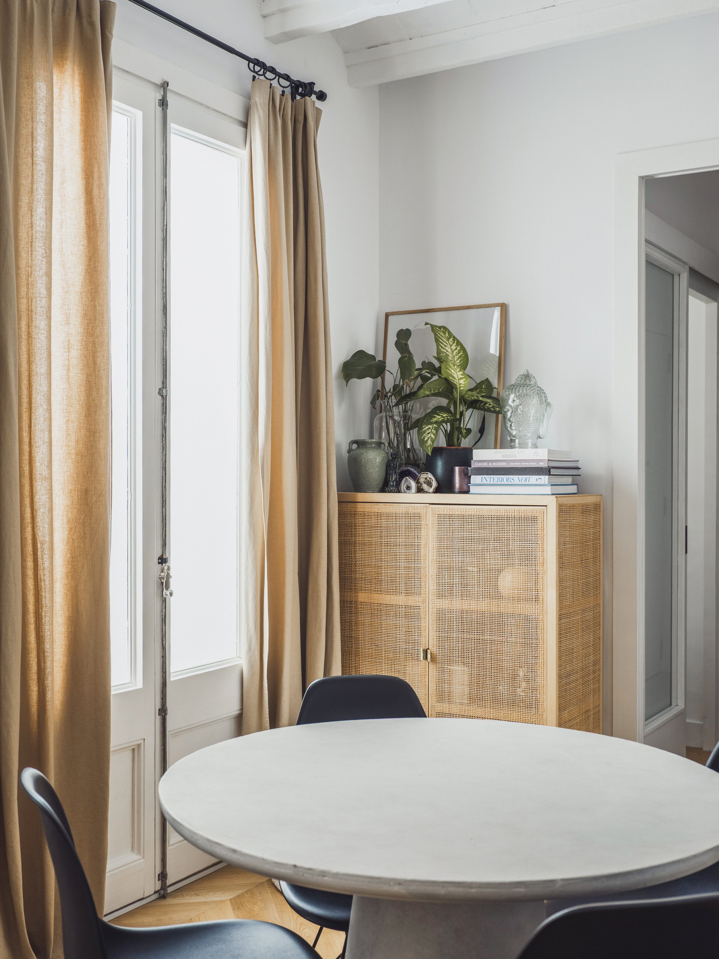 A white round table with black chairs around it, a cabinet with plants, books, and decorative objects, a mirror leaning against the wall, beige curtains on a large window, and a doorway leading to another room.