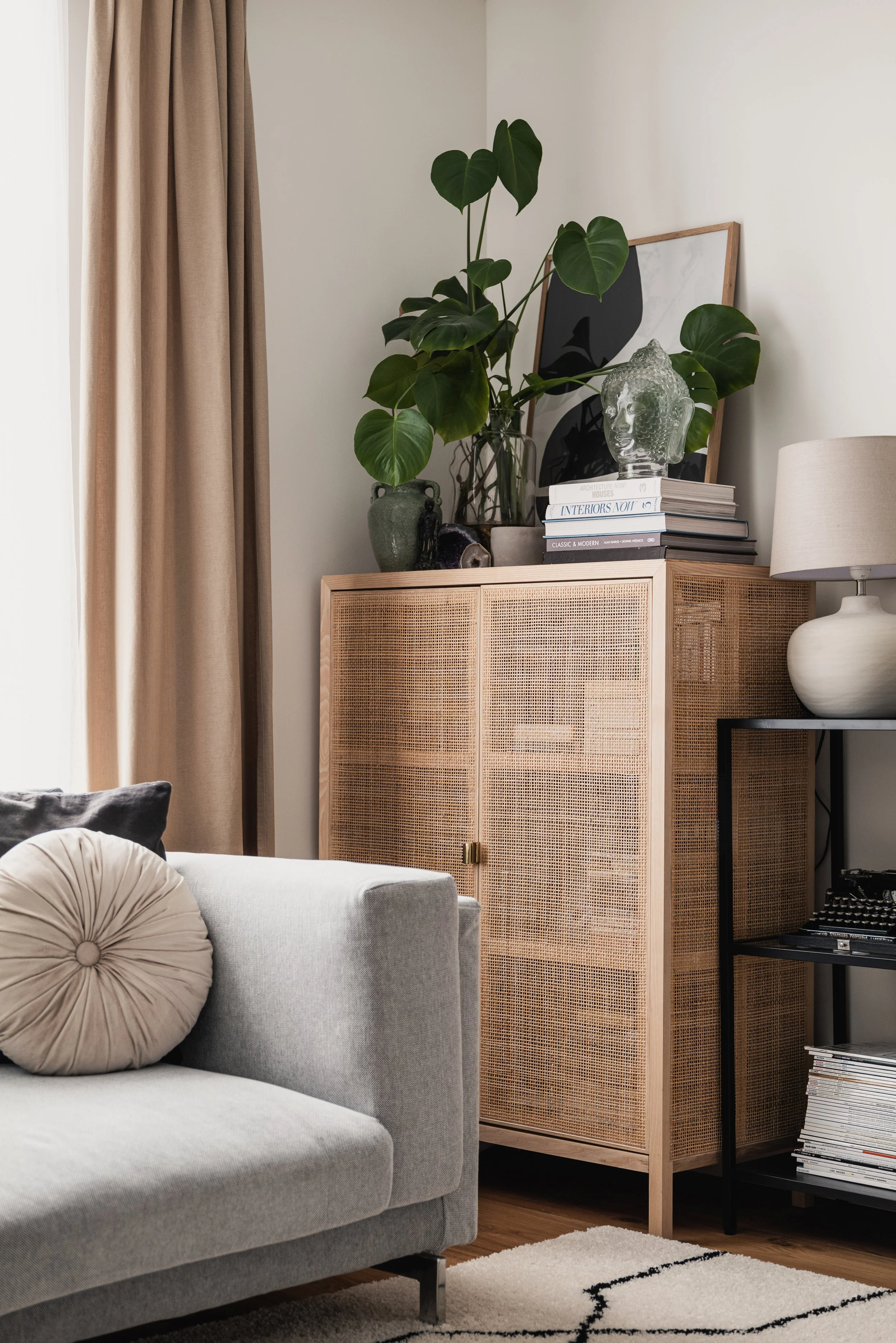 Living room corner with beige curtains, a light gray sofa with a round textured pillow, a rattan cabinet decorated with plants, books, a glass bust, and framed artwork, and a black side table with a beige lamp and magazines.