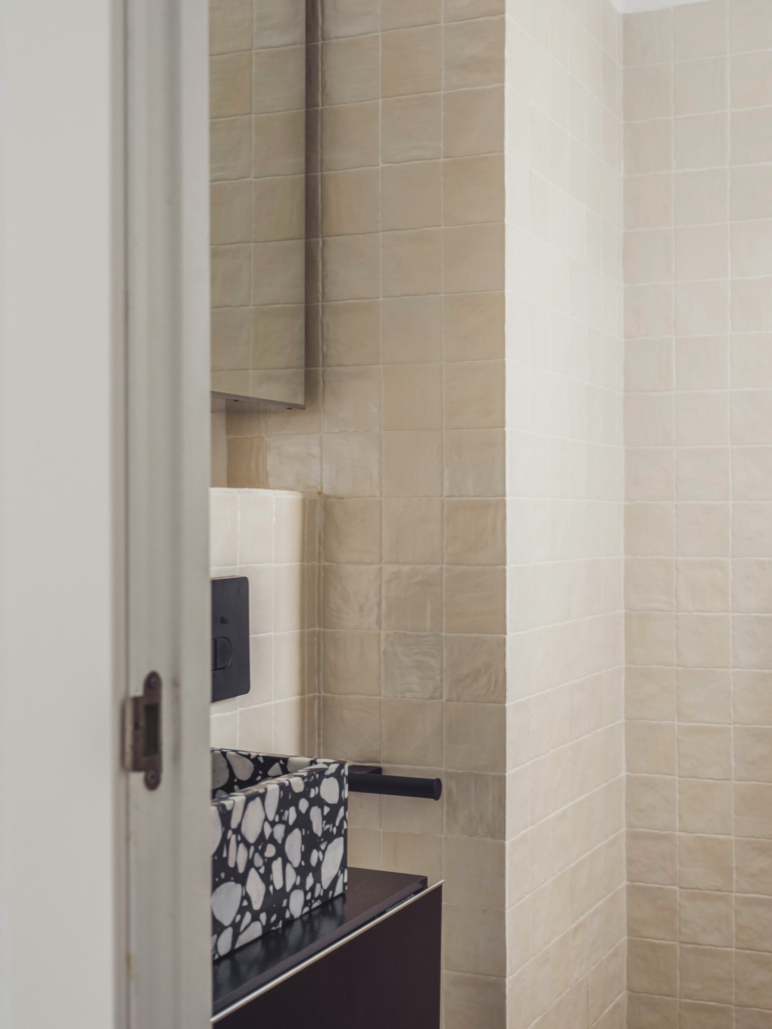 A bathroom with beige tiled walls, a small mirror on the wall, a black towel with white abstract patterns hanging from a black rack, and a black wall switch or outlet visible near the towel.