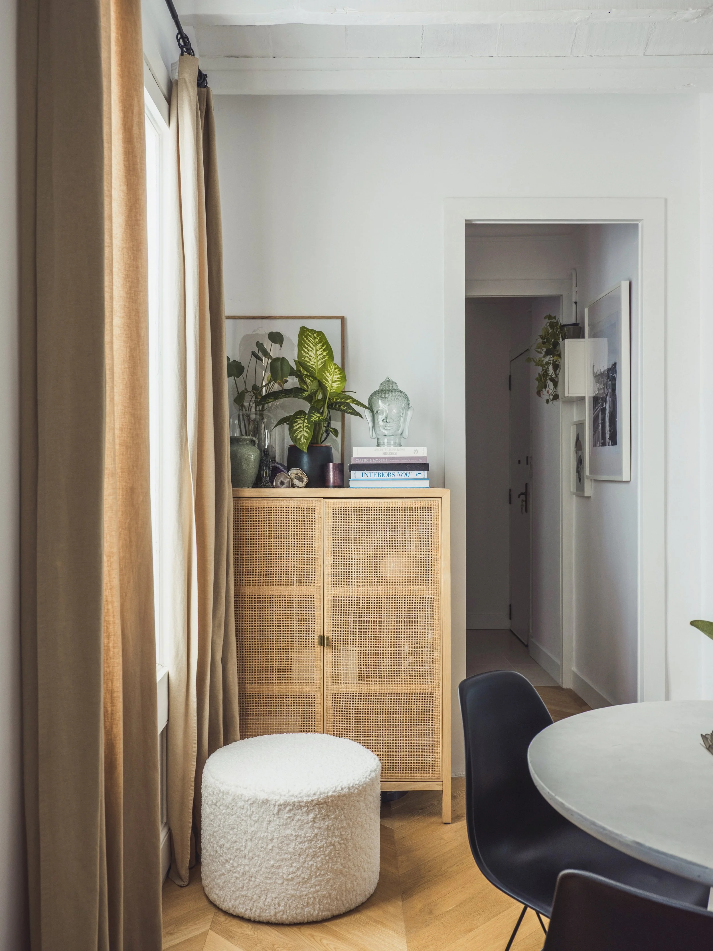 Interior of a home with beige curtains, a white textured round ottoman, a wooden cabinet with rattan doors, potted plants, and a glass Buddha head sculpture on top. There is a doorway and a glimpse of a staircase in the background.