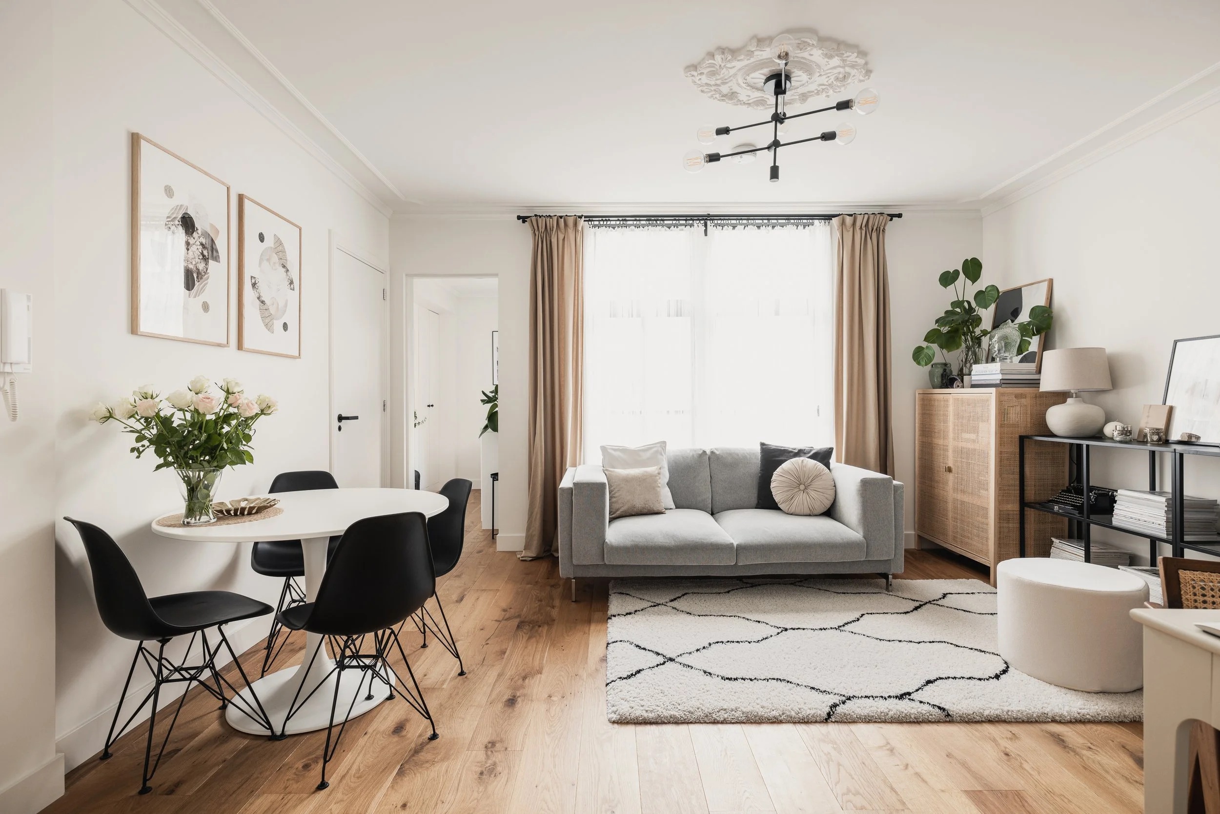Living room with a gray sofa, cream curtains, a patterned black and white rug, a white oval dining table with four black chairs, and various decorative plants and artwork.
