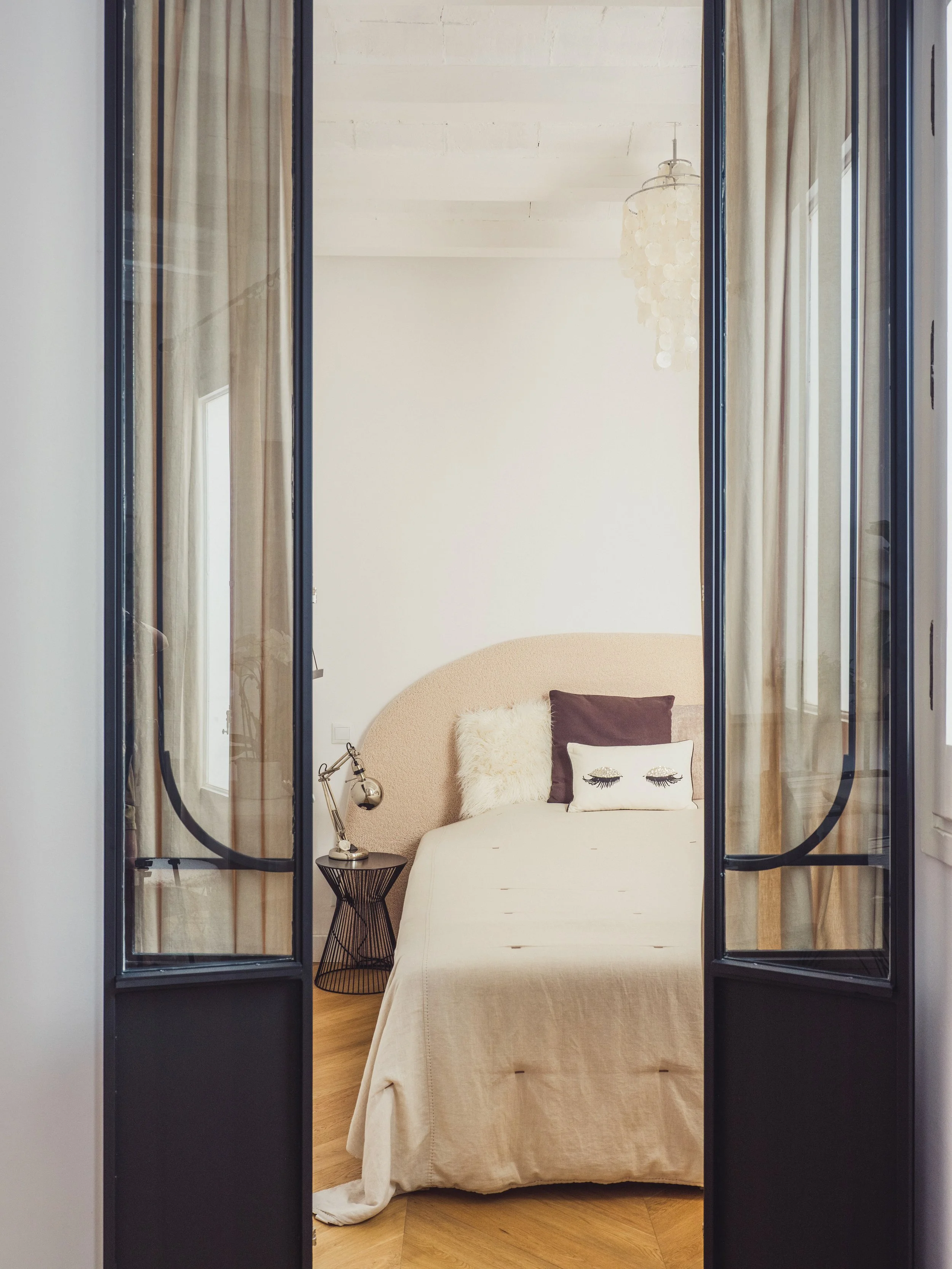 A bedroom viewed through black-framed glass doors. The bed has a beige linen cover, decorated with pillows including one with closed eyes design, and is flanked by a black wire side table with a silver desk lamp. A hanging chandelier and beige curtai