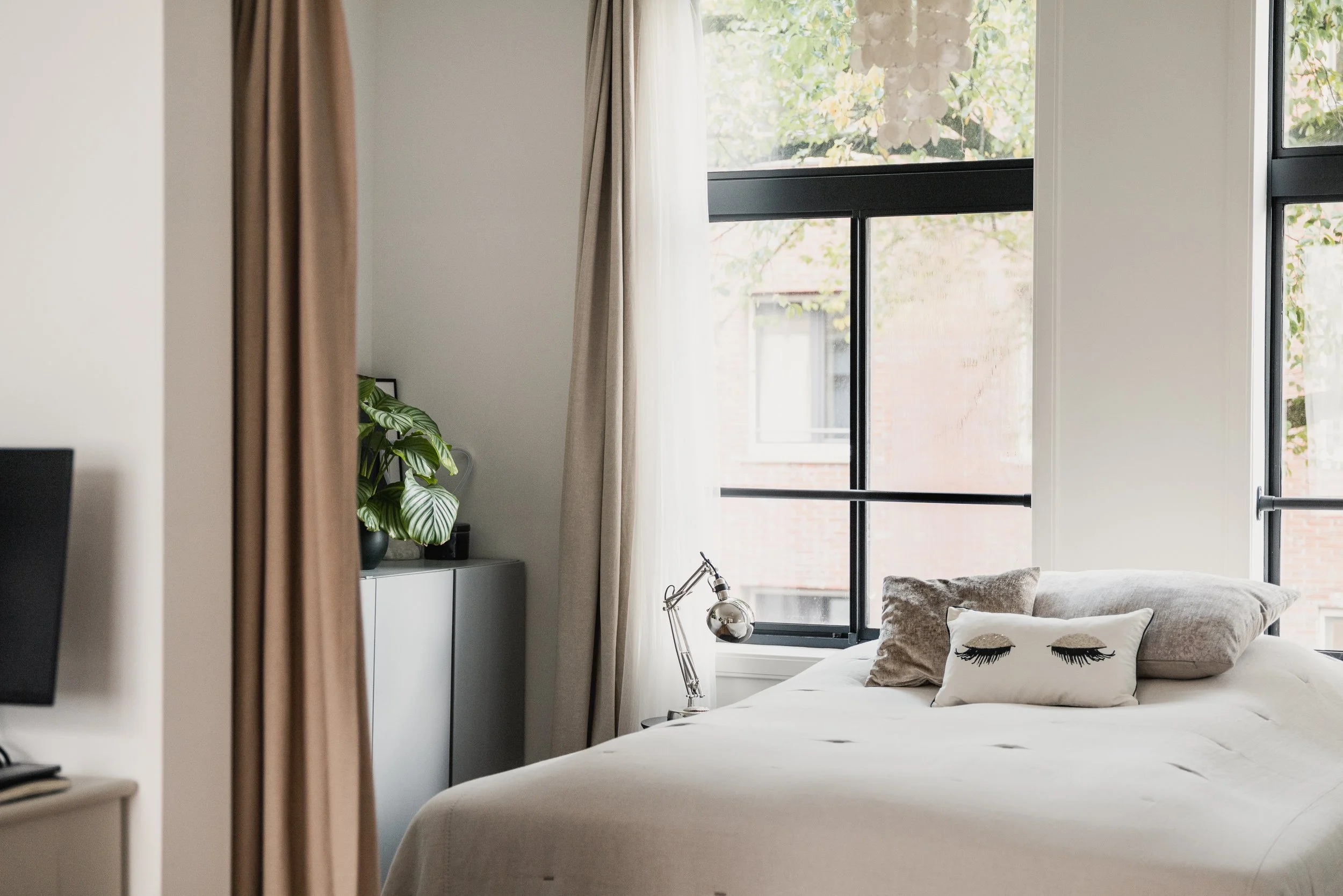 A neatly made bed with decorative pillows, including one with closed eyelash designs, in front of large windows with beige curtains, and a desk lamp on a side table near a window with outdoor trees visible.