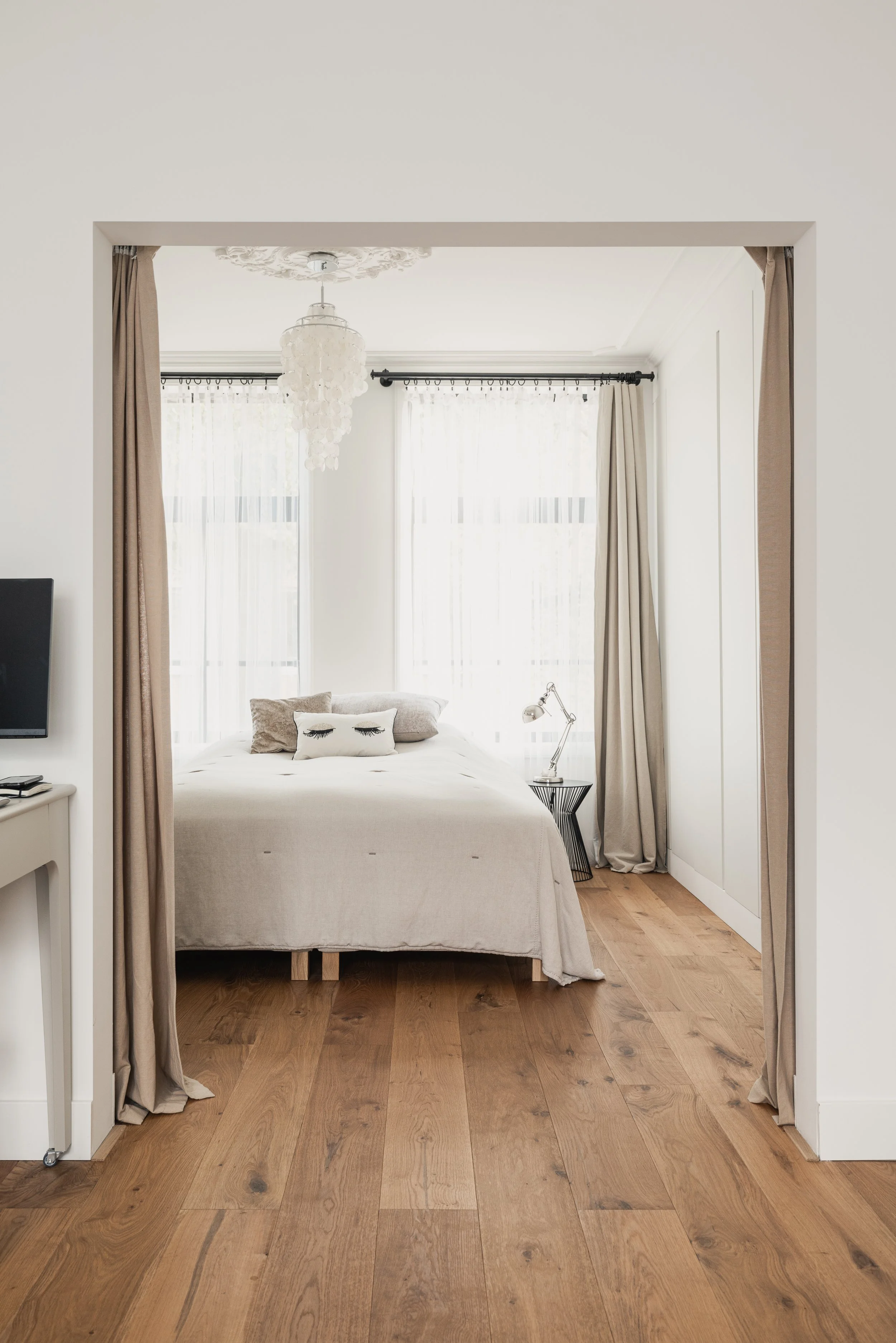 Cozy bedroom with beige curtains, white bedding, pillows with eyelash designs, wooden floor, small bedside table with adjustable lamp, and large windows with sheer white curtains.
