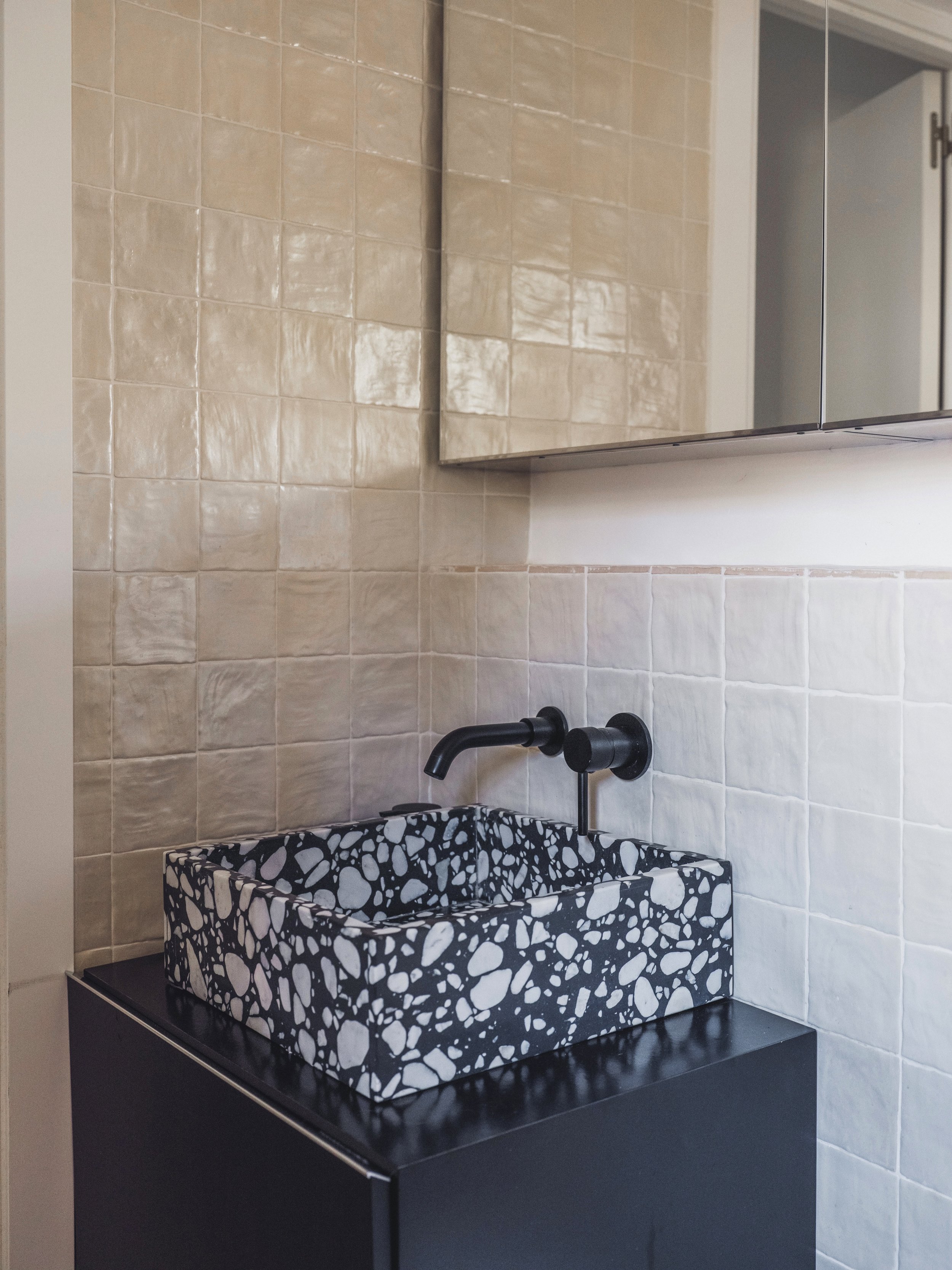 Kitchen with beige tiled walls, black countertop, black and white terrazzo style hand wash basin, black wall-mounted faucet, mirrored cabinet above