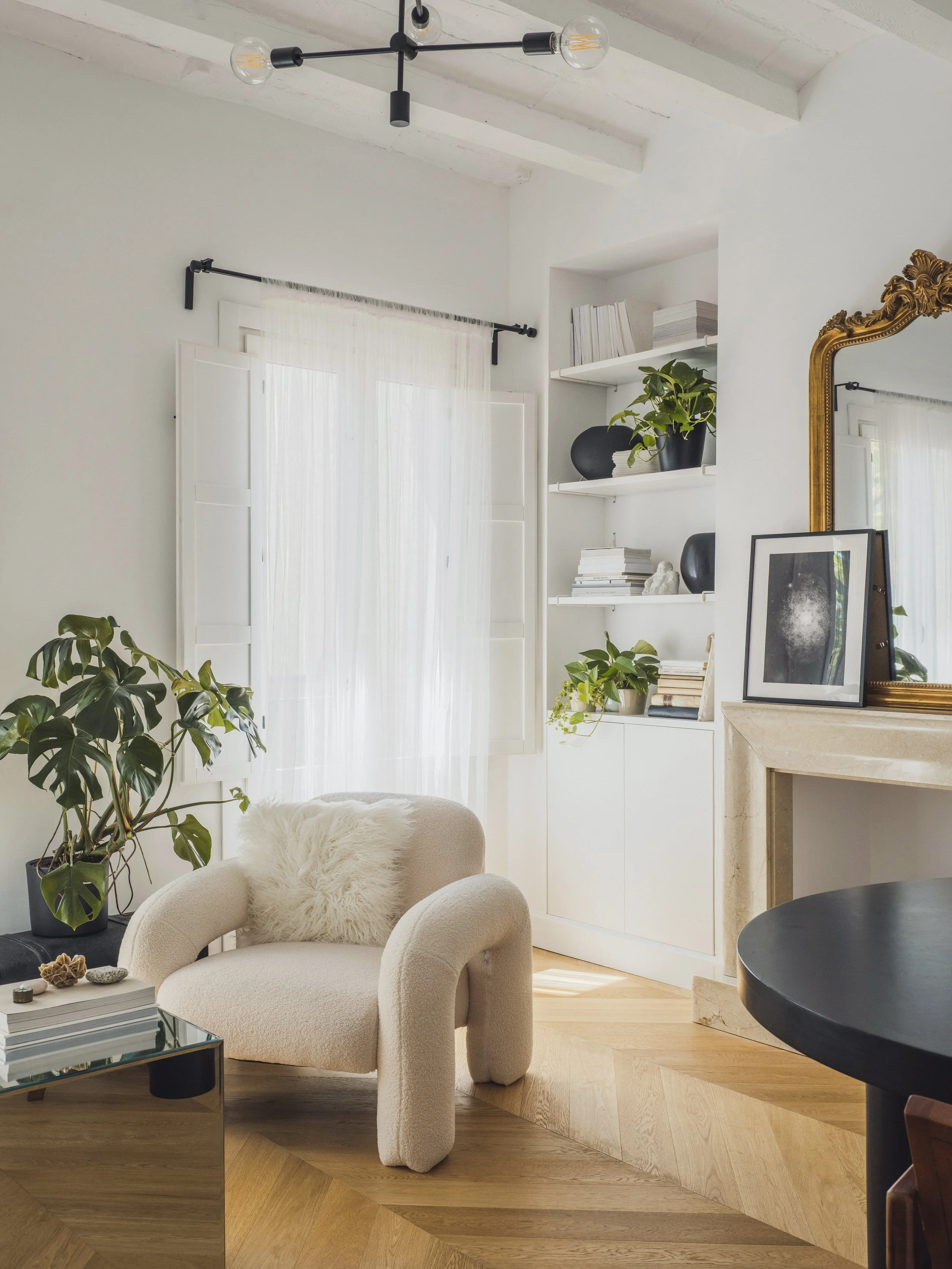 A living room with white walls and a ceiling with exposed beams. There is a white curtain covering a window, a beige armchair with a fluffy pillow, and a black table with some decorative stones. Shelves hold books, a black vase, and a potted plant. A