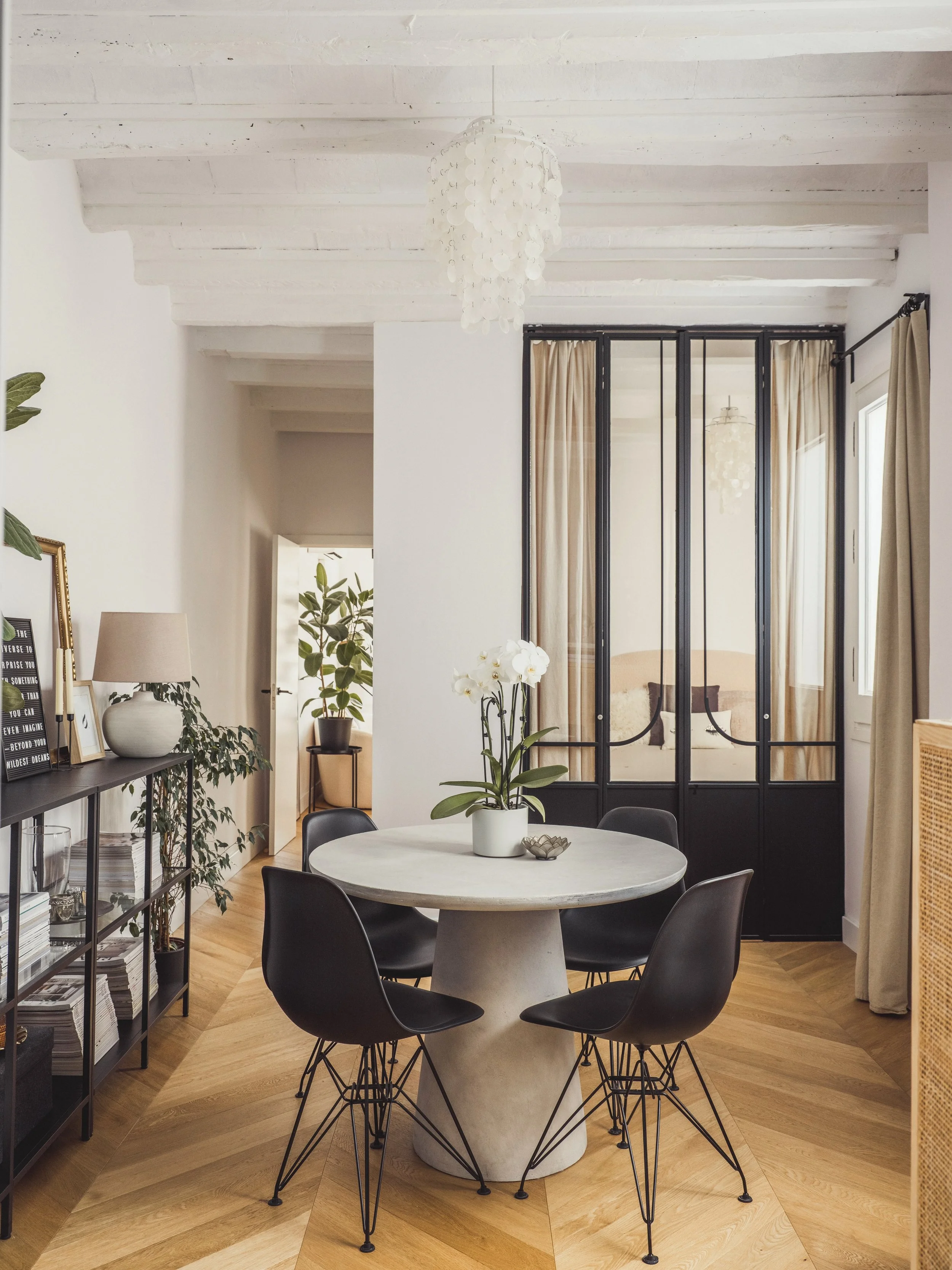 A modern dining area with a round white marble table surrounded by six black chairs with wire bases. There is a potted white orchid in the center of the table, and the room features a light wood herringbone floor. On the left side, a black shelving u