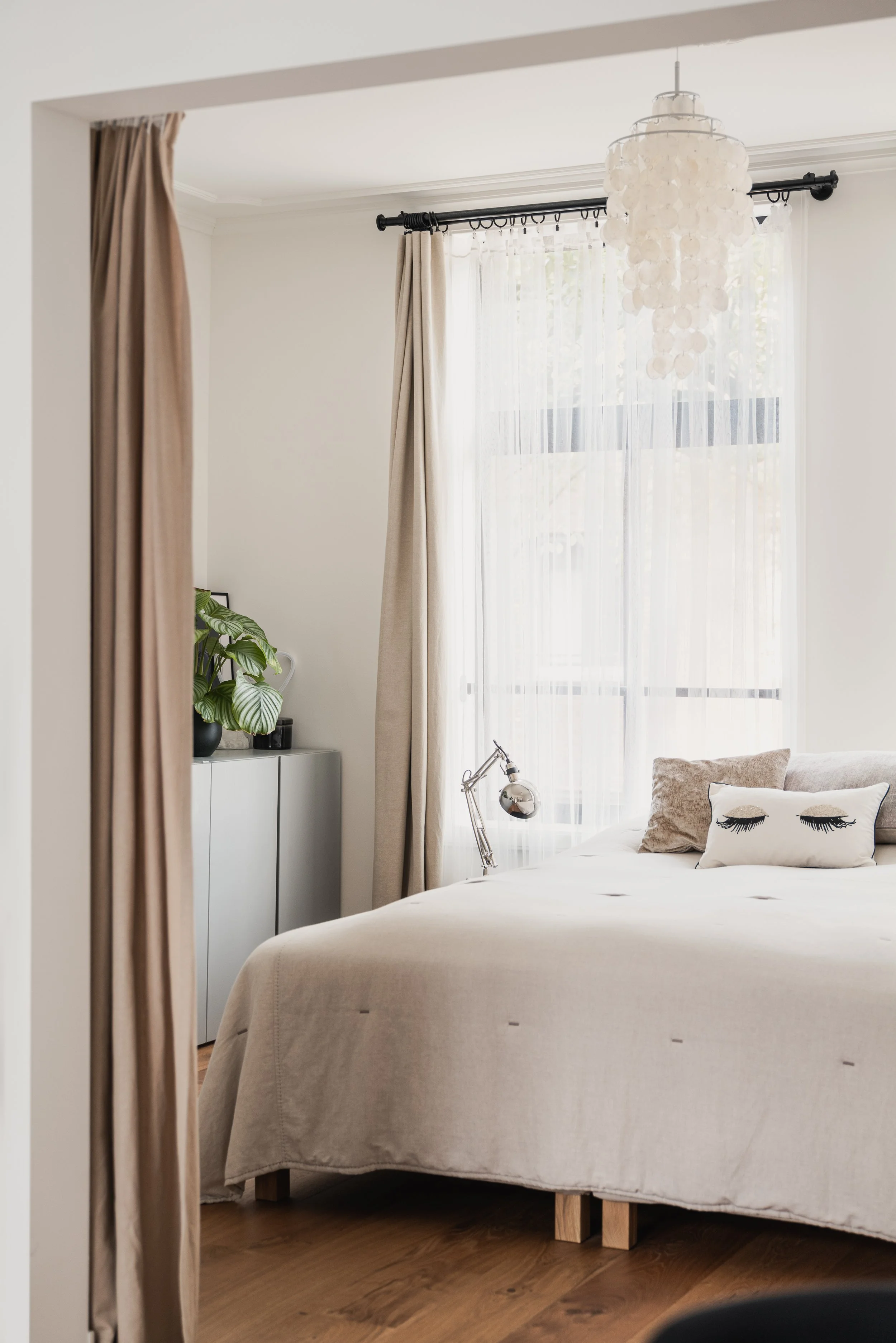 Bright bedroom with large window, beige curtains, white bedspread, decorative pillows with eyelash designs, small silver desk lamp, and a white cabinet with a green houseplant.