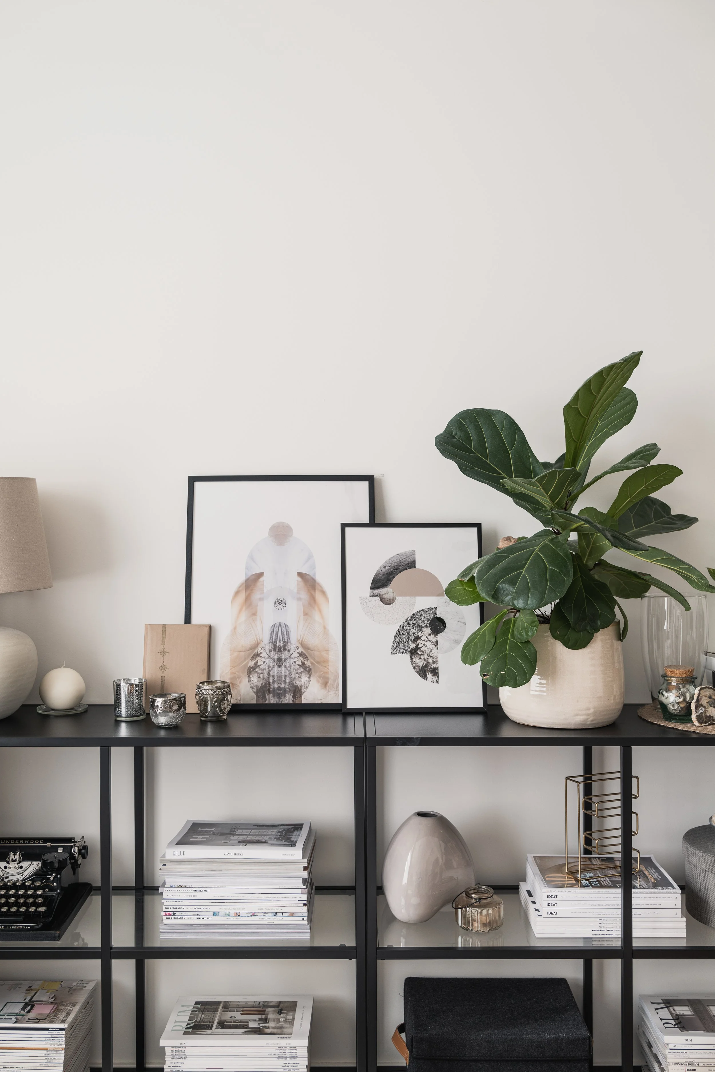 Modern black shelving unit with books, framed art, decorative items, and a large potted fiddle leaf fig plant against a white wall.