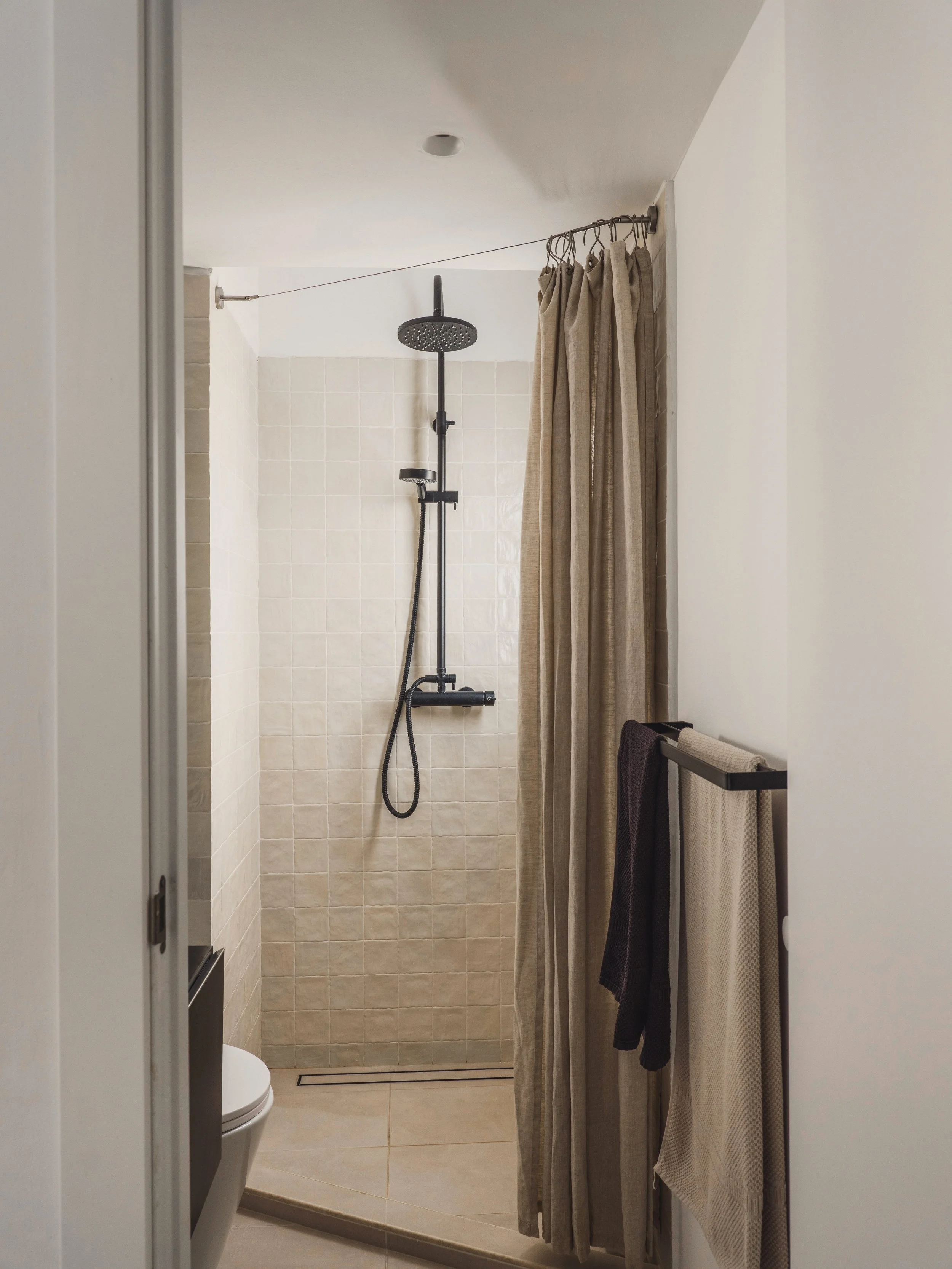 View of a minimalist shower area with a black rainfall showerhead, beige tiled wall, beige shower curtain, folded towels hanging on a towel rack, and part of a toilet in a small bathroom.