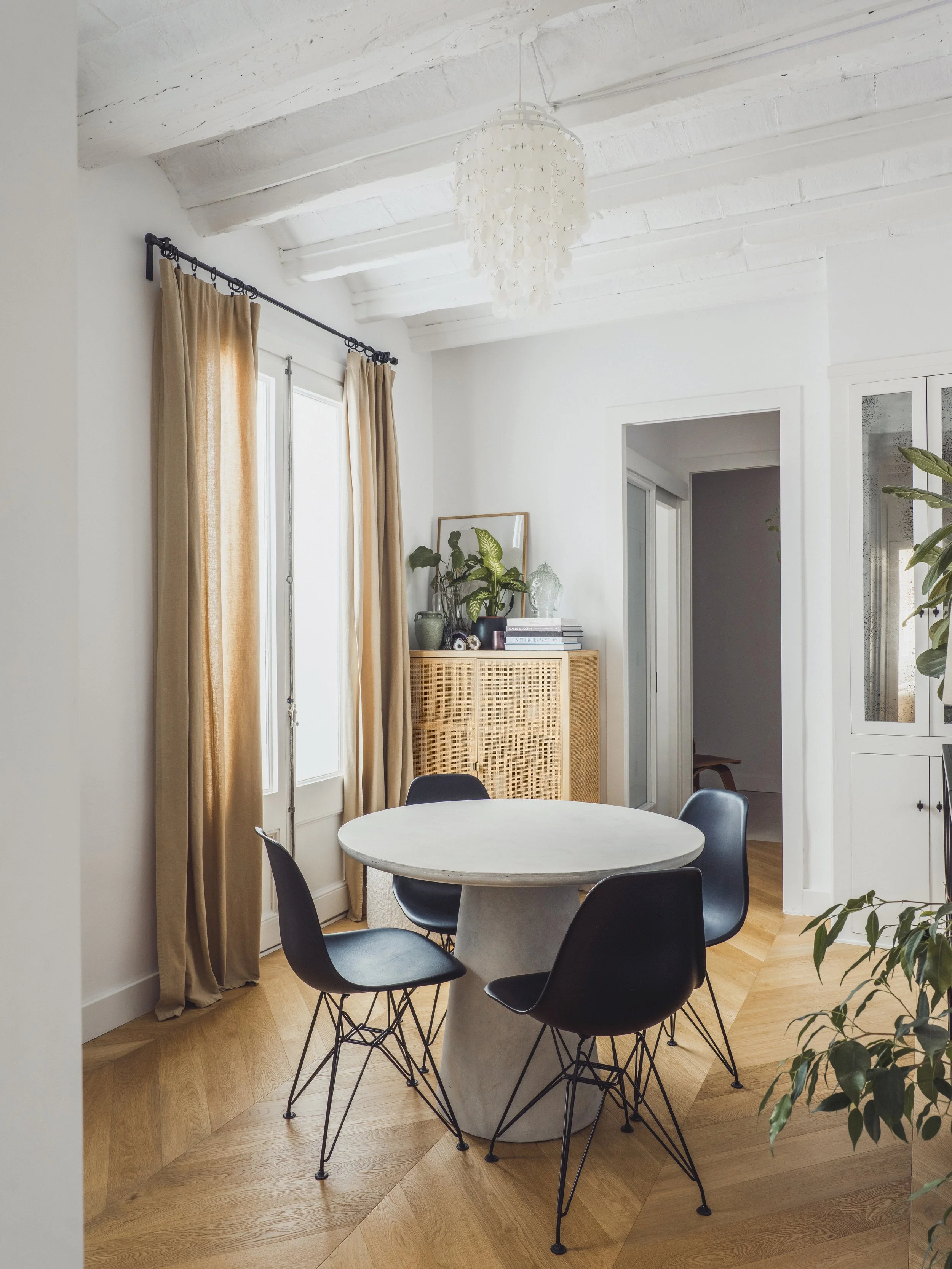A dining area with a round marble table and four black chairs with wire bases on a wooden floor, light-colored curtains on windows, a white ceiling, a ceiling lamp, a corner cabinet with plants and books, and a doorway leading to another room.