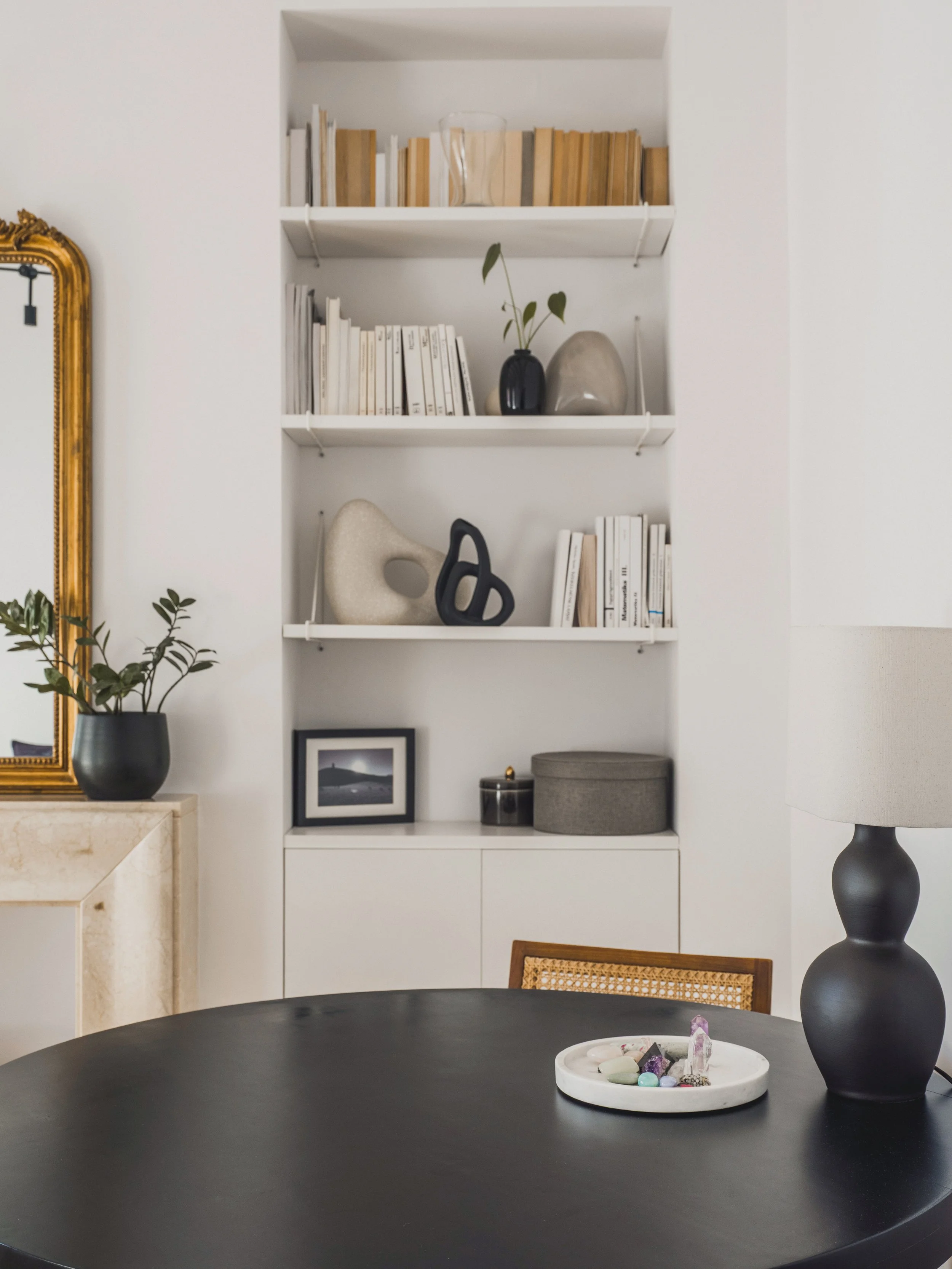A black round table with a white decorative tray containing crystals and stones. To the right, a large dark gray ceramic vase and a smaller gray box. In the background, a white shelving unit with books, vases, and decorative objects, a framed black-a