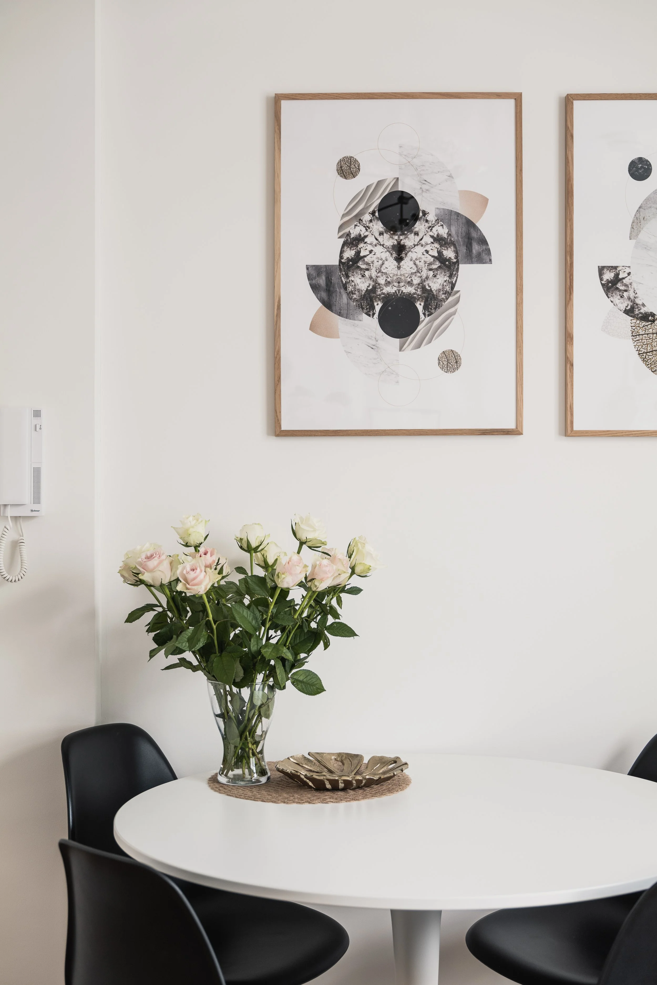 White dining table with a glass vase filled with pale pink roses and green leaves, surrounded by black chairs. Wall art featuring abstract black, white, and beige shapes hangs on a white wall.