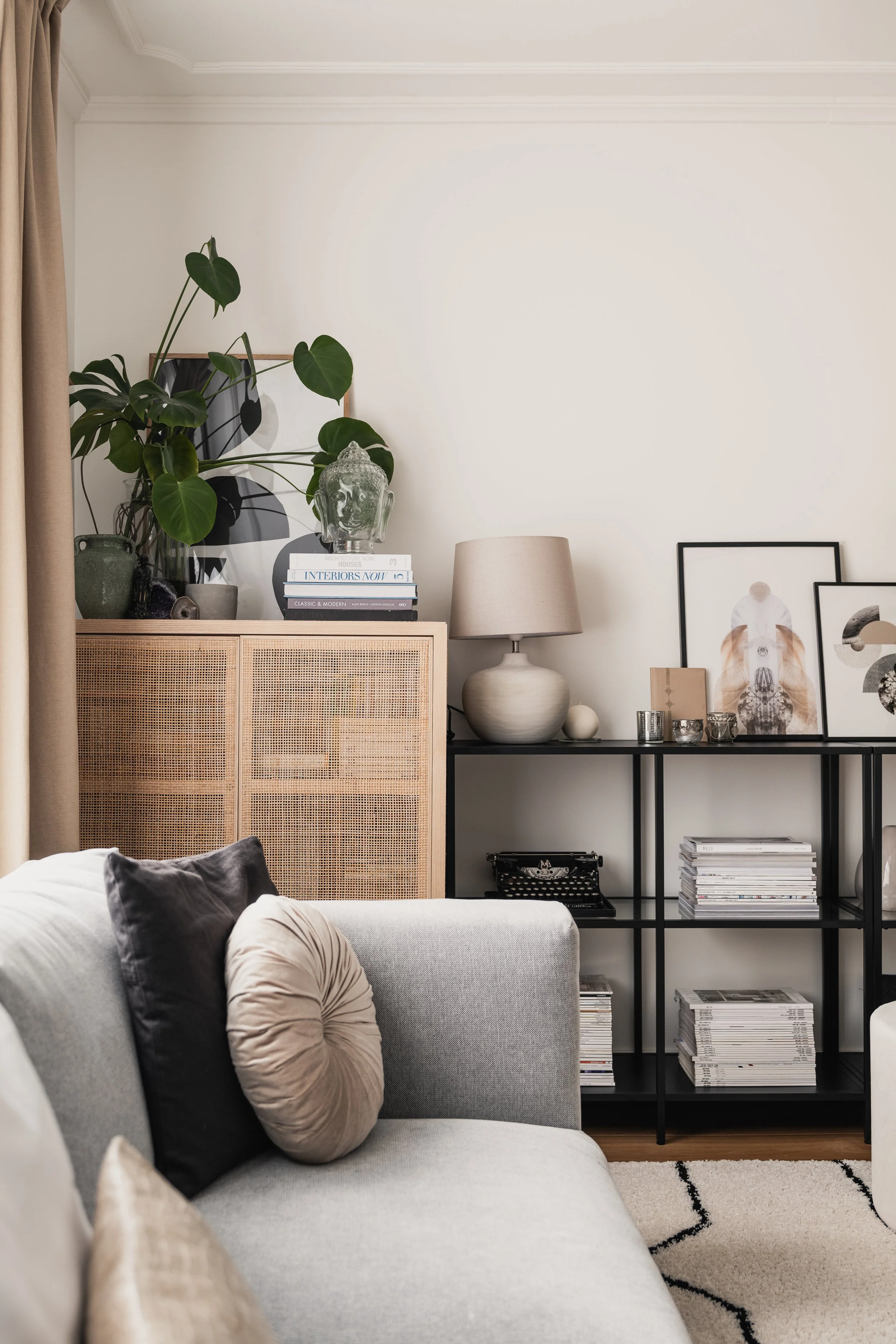Living room corner with a beige couch, black and beige pillows, a wooden cabinet, and a black shelving unit with artwork, books, and decorative objects.