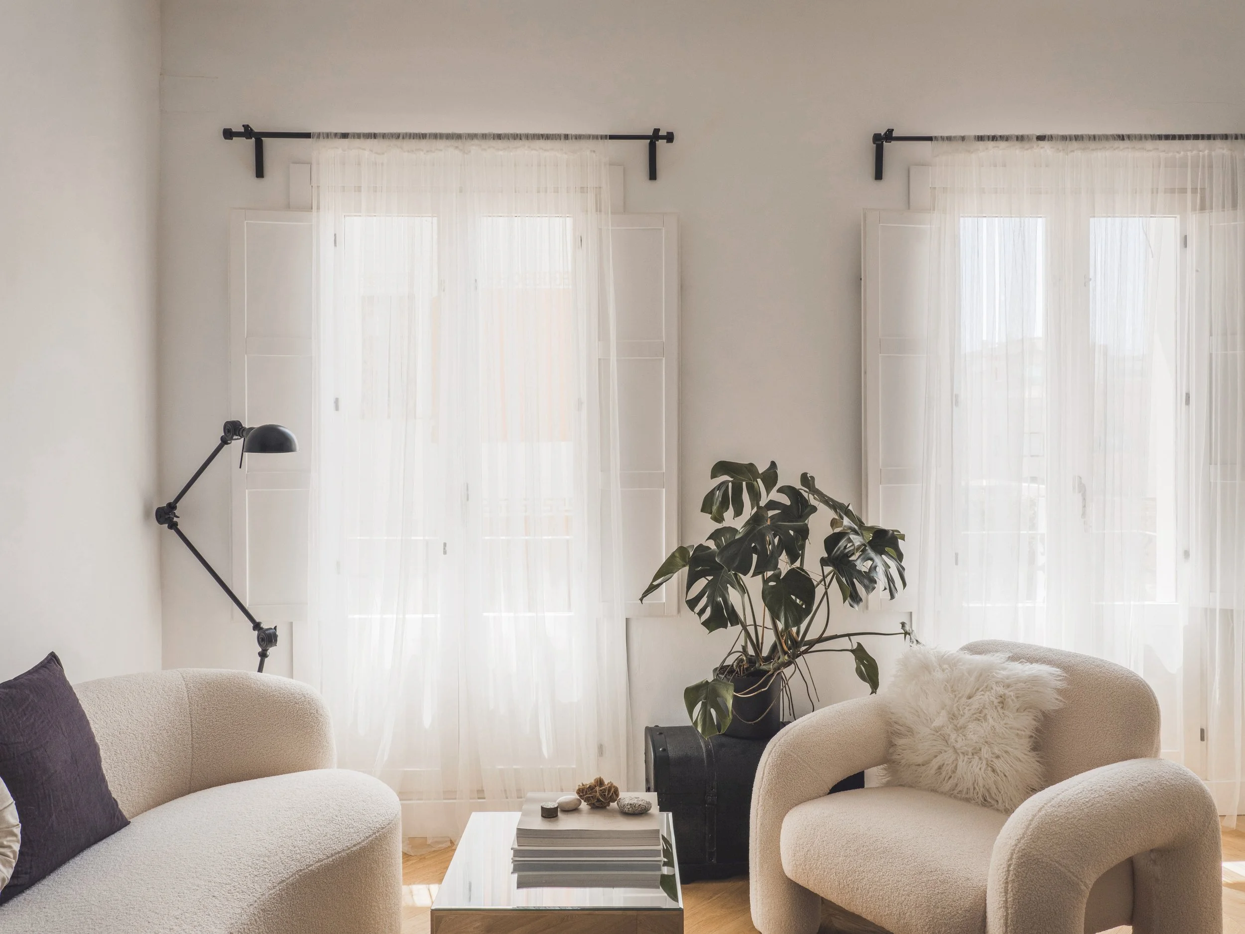 Living room with white curtains on large windows, beige seating, a black floor lamp, a potted plant, and a coffee table with books and decorative items.
