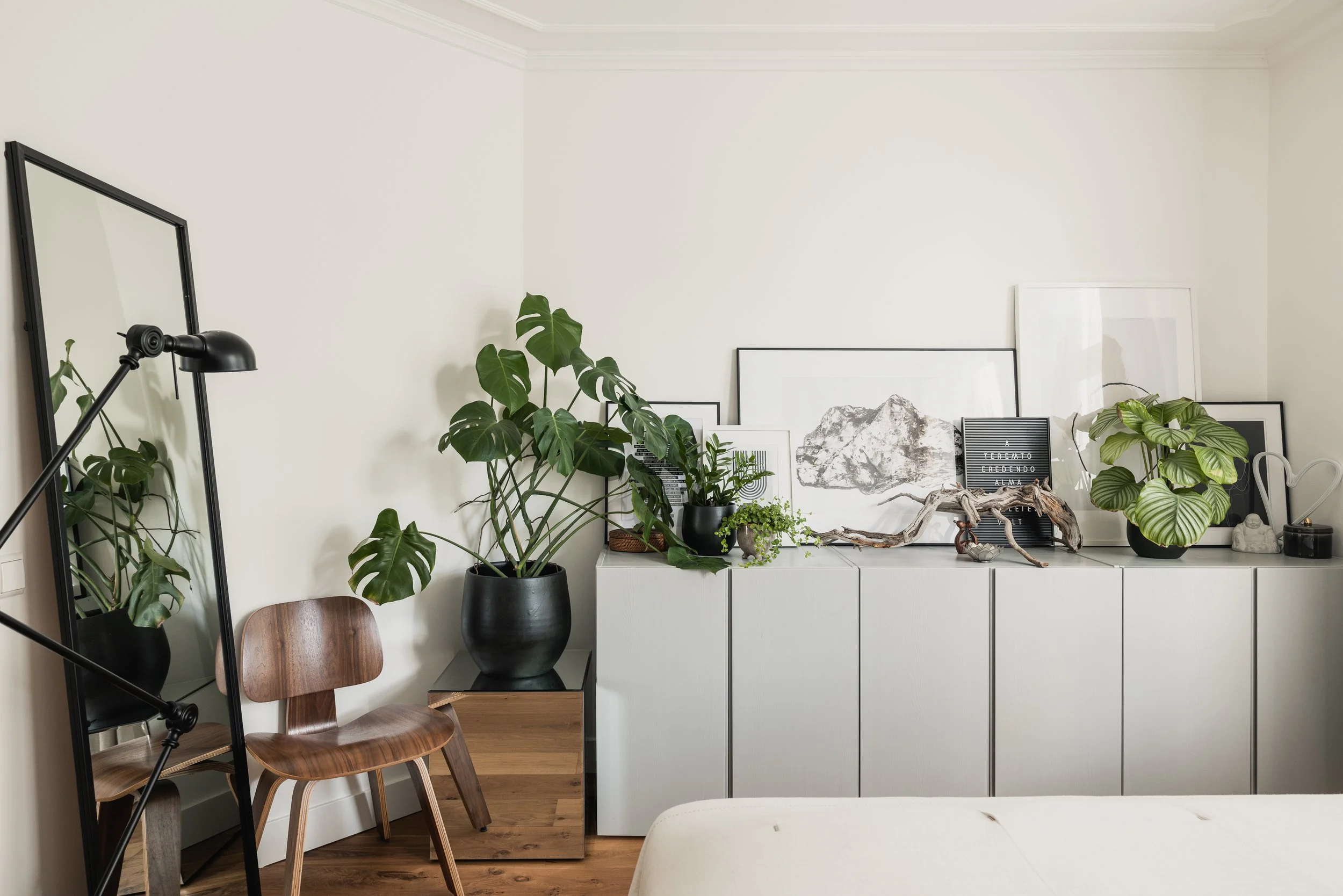 Decorative bedroom corner with black-framed mirror, wooden chair, large indoor plants, framed art, and a white cabinet.