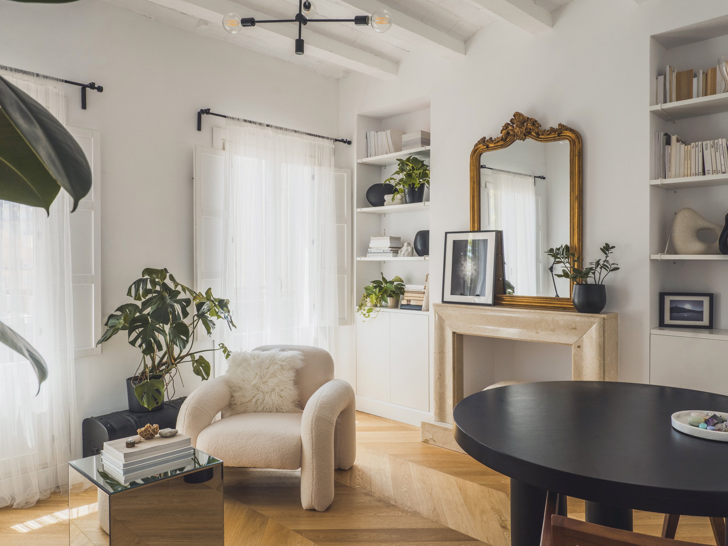 A cozy living room with a plush beige armchair, a glass coffee table with books and decorative objects, large windows with sheer curtains, and a built-in bookshelf with books and plants. A large ornate gold mirror hangs on the wall above a fireplace,
