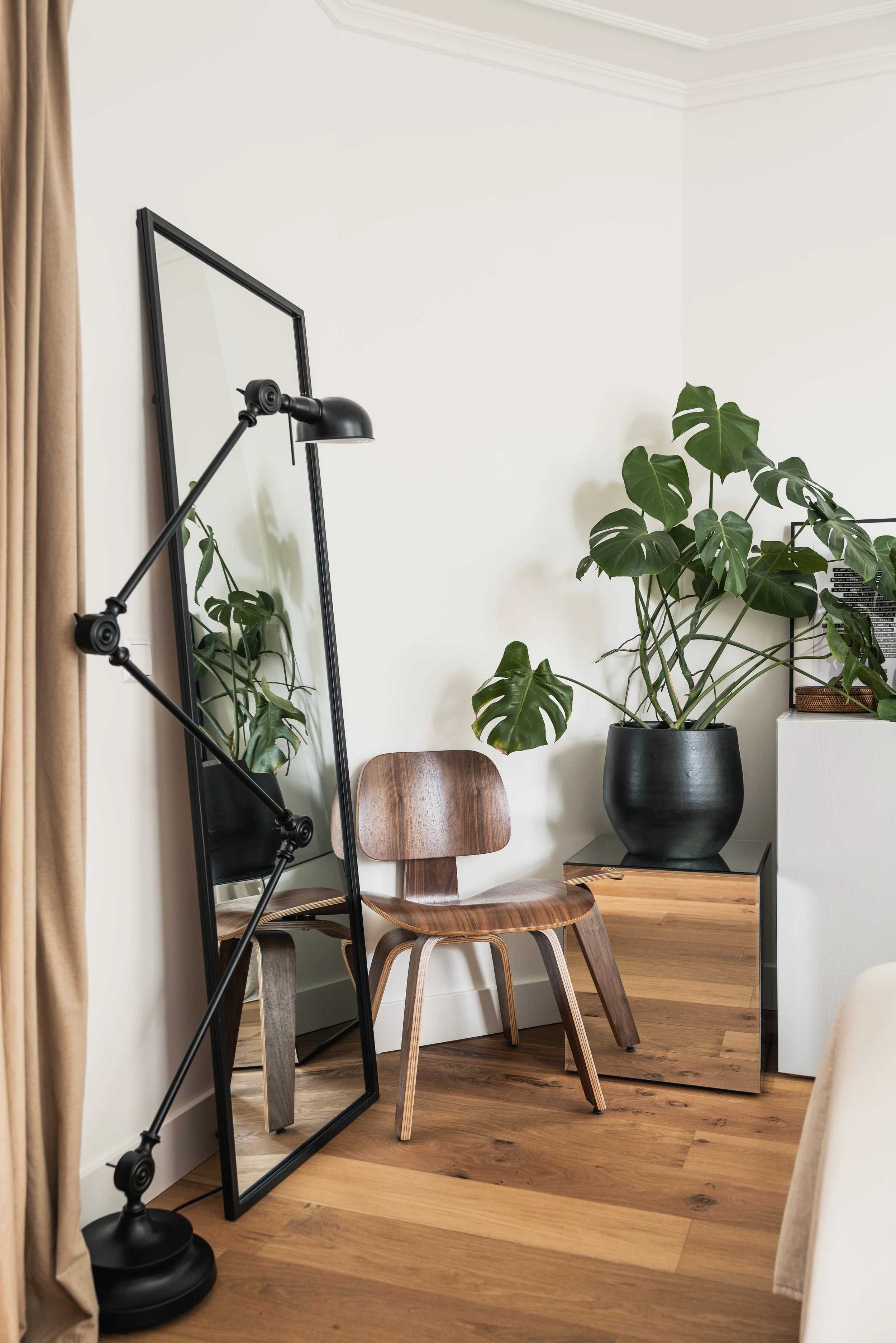 A room corner with a large mirror, a wooden chair, and a potted monstera plant on a wooden side table.