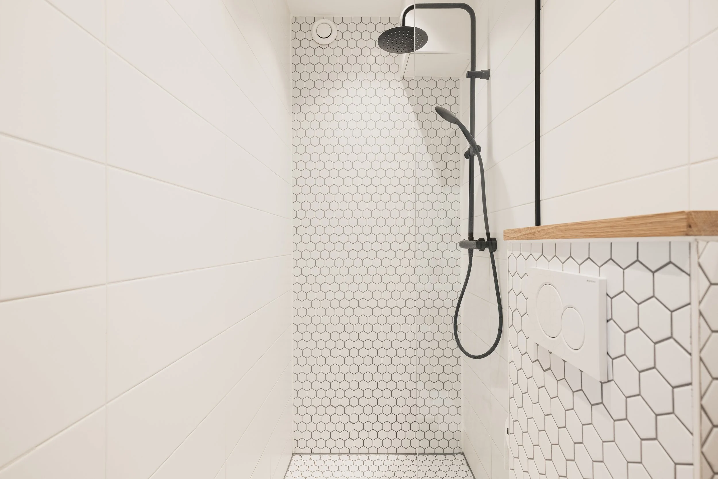 White tiled walk-in shower with black rainfall and handheld showerheads, hexagon mosaic tiles on the back wall, and a wooden shelf on the right side.