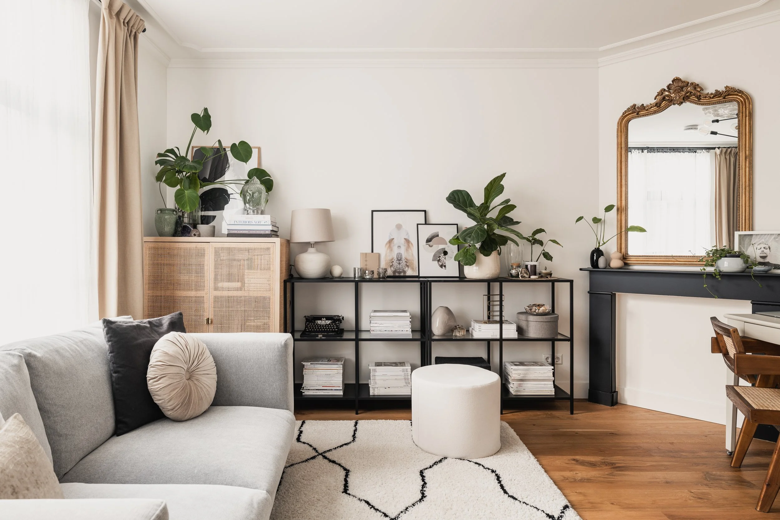 Living room with a light gray sofa, black metal bookshelf with books and decorative objects, large green plants, a white ottoman, a mirror with a gold frame, and a black fireplace mantel decorated with plants and art.
