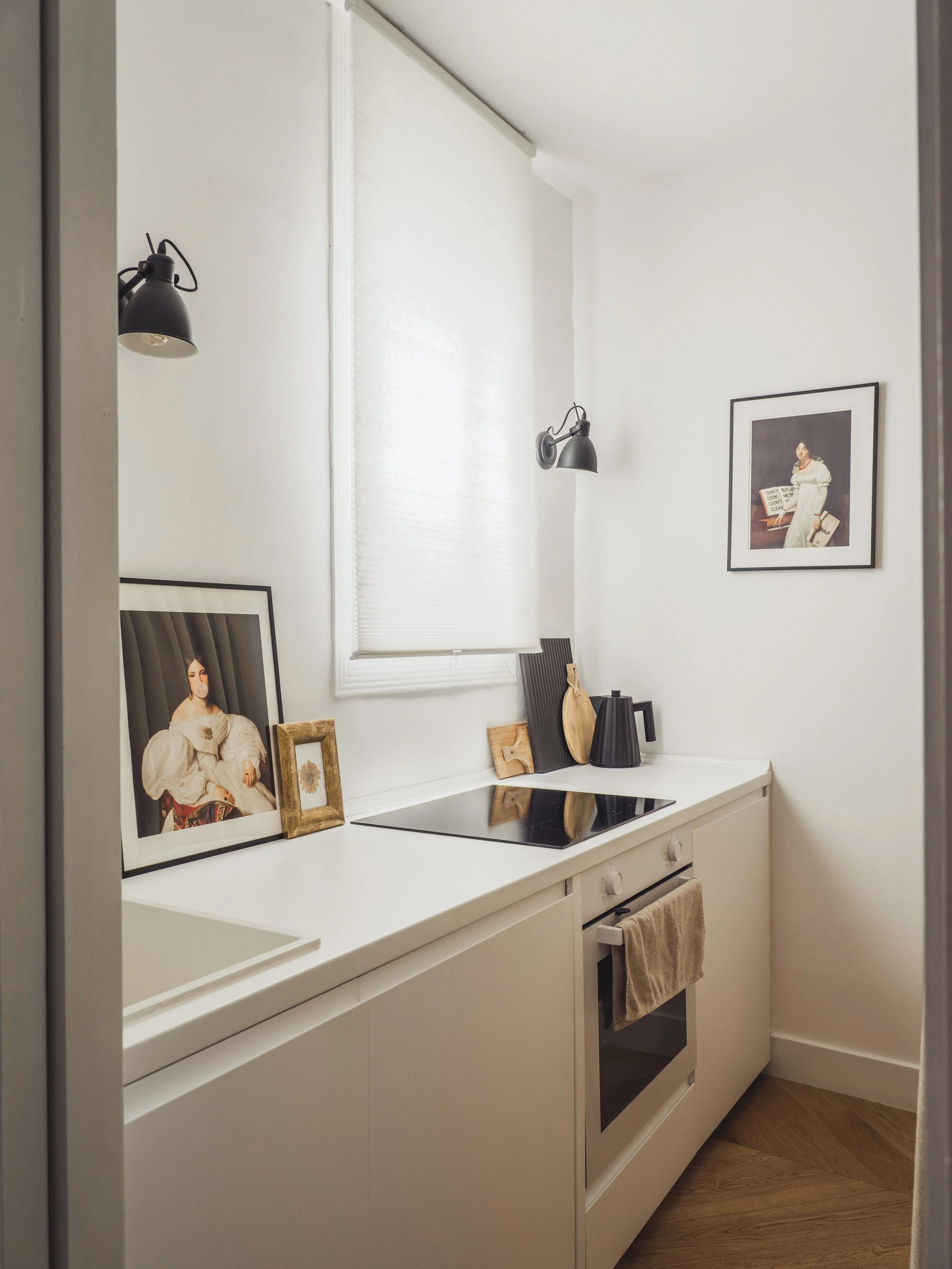 A small kitchen countertop with framed pictures, a black pitcher, and wooden cutting boards. There are two black wall-mounted lamps above, and a window with blinds. A beige towel hangs from the oven handle.