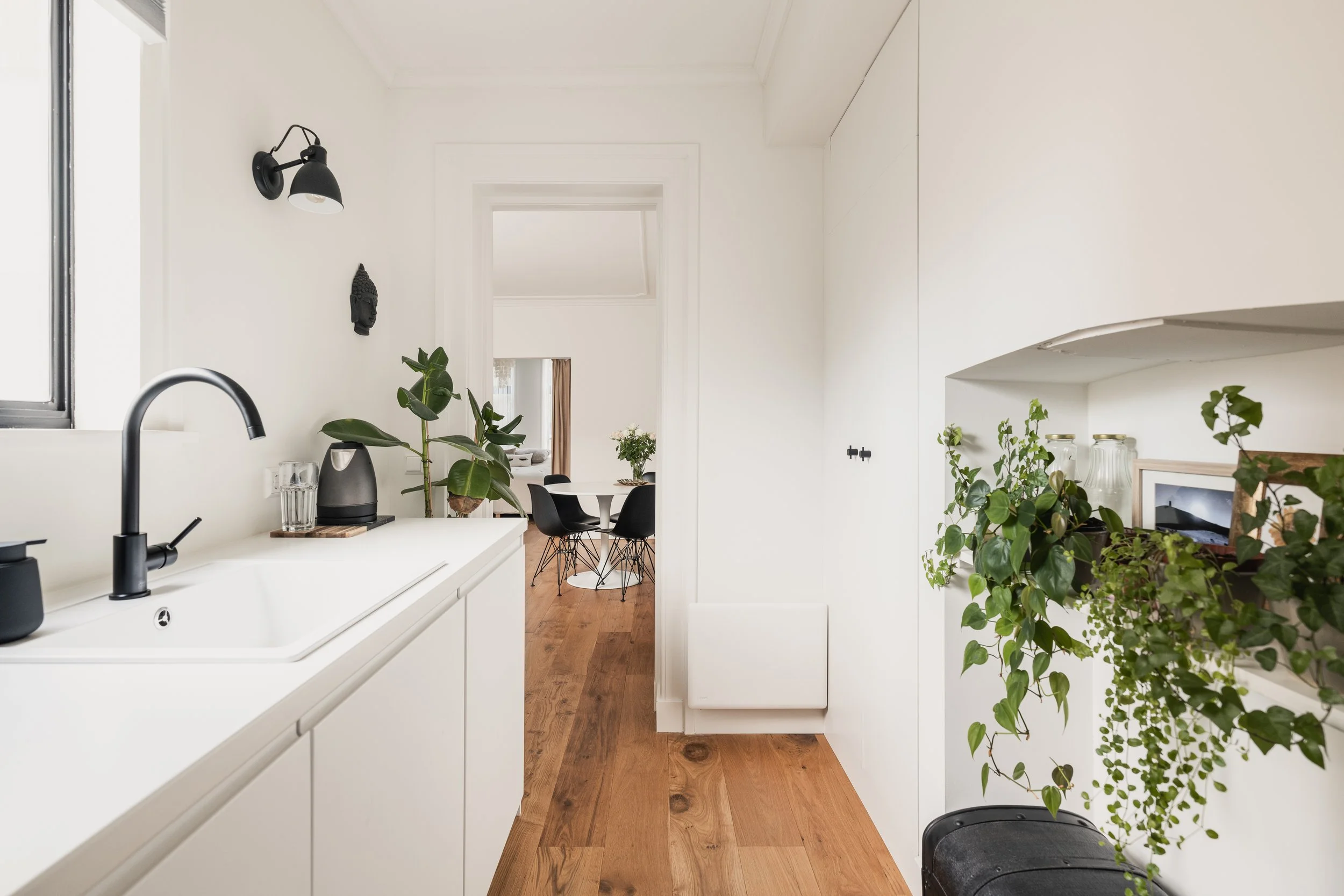 Modern kitchen with white cabinets, black fixtures, wooden flooring, and green plants, leading to a dining area with a black table and chairs.