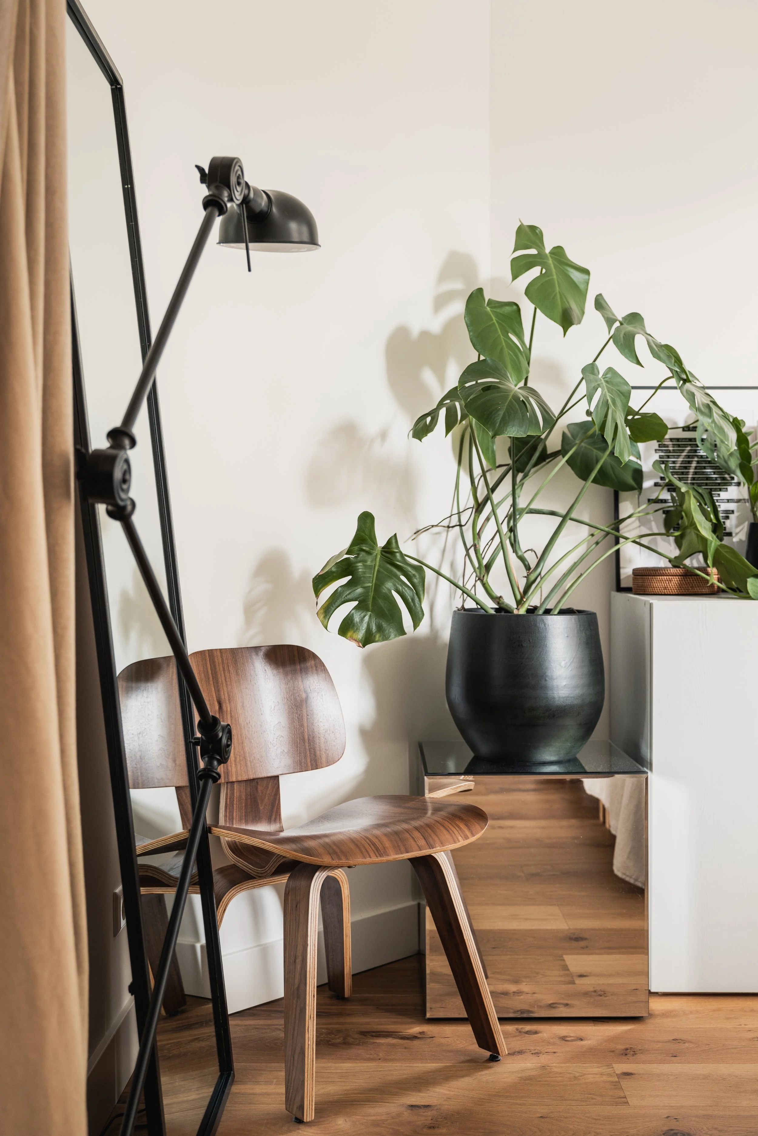 A wooden chair, a large potted monstera plant, a metallic side table, a black floor lamp, beige curtains, and a white cabinet with a smaller plant and a basket on top.