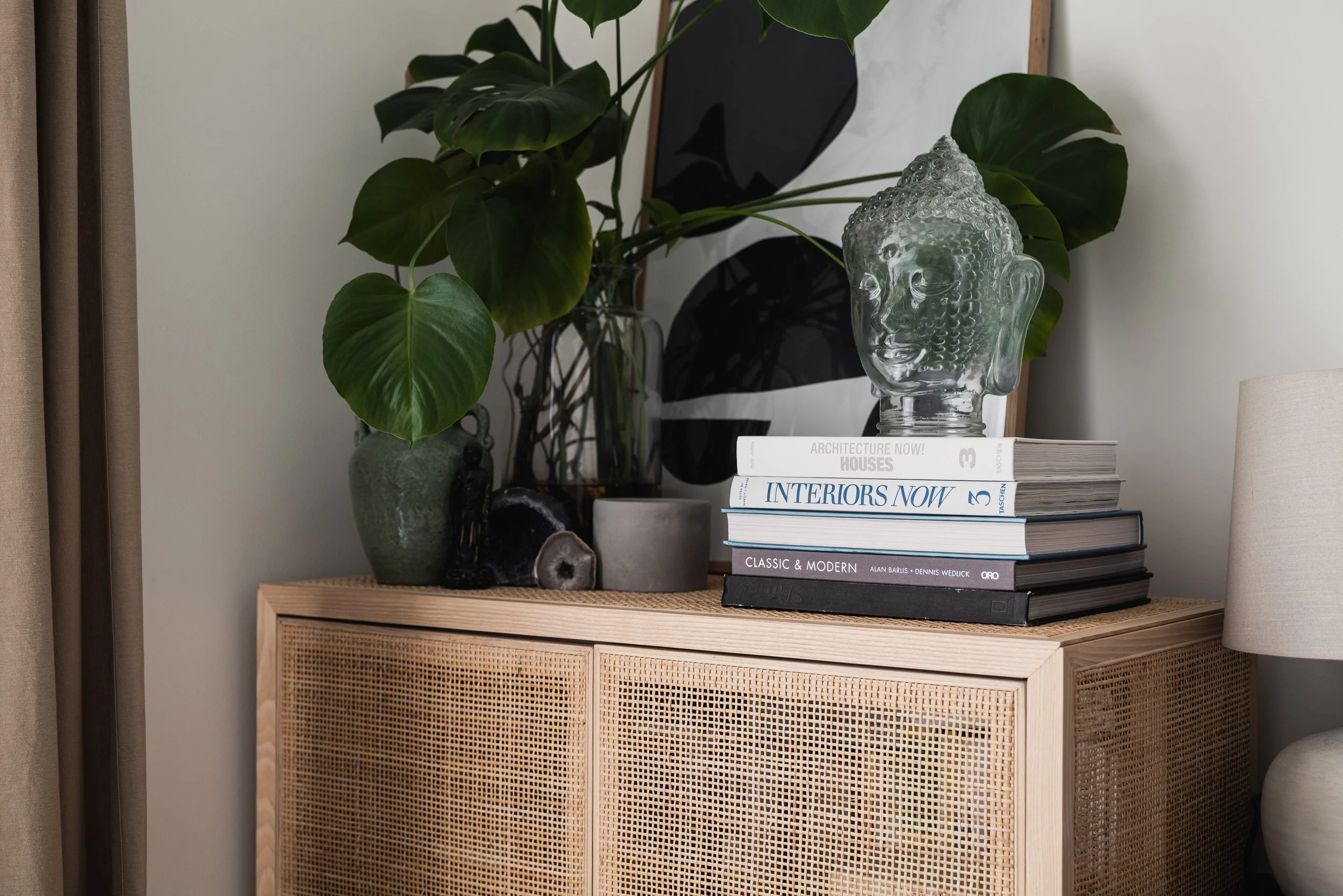 A wooden cabinet holding decorative items including a large green leafy plant in a ceramic pot, a Buddha head sculpture, and stacked books on interior design and architecture, with a framed black and white abstract art piece behind them.