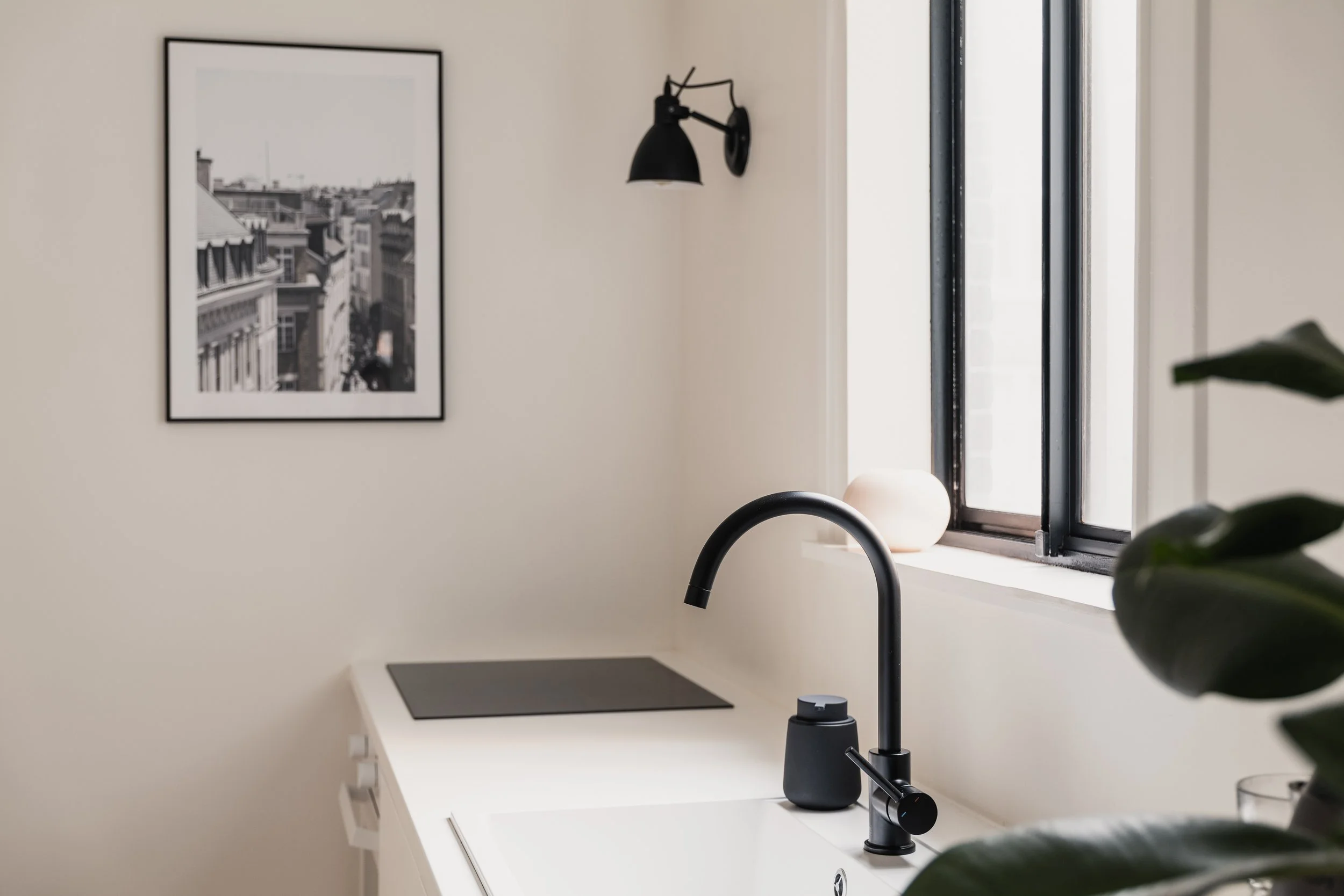 A modern kitchen corner with a black faucet, a white countertop, a window, a black and white framed cityscape photograph on the beige wall, a black wall sconce, a white decorative object on the windowsill, and some green leaves in the foreground.