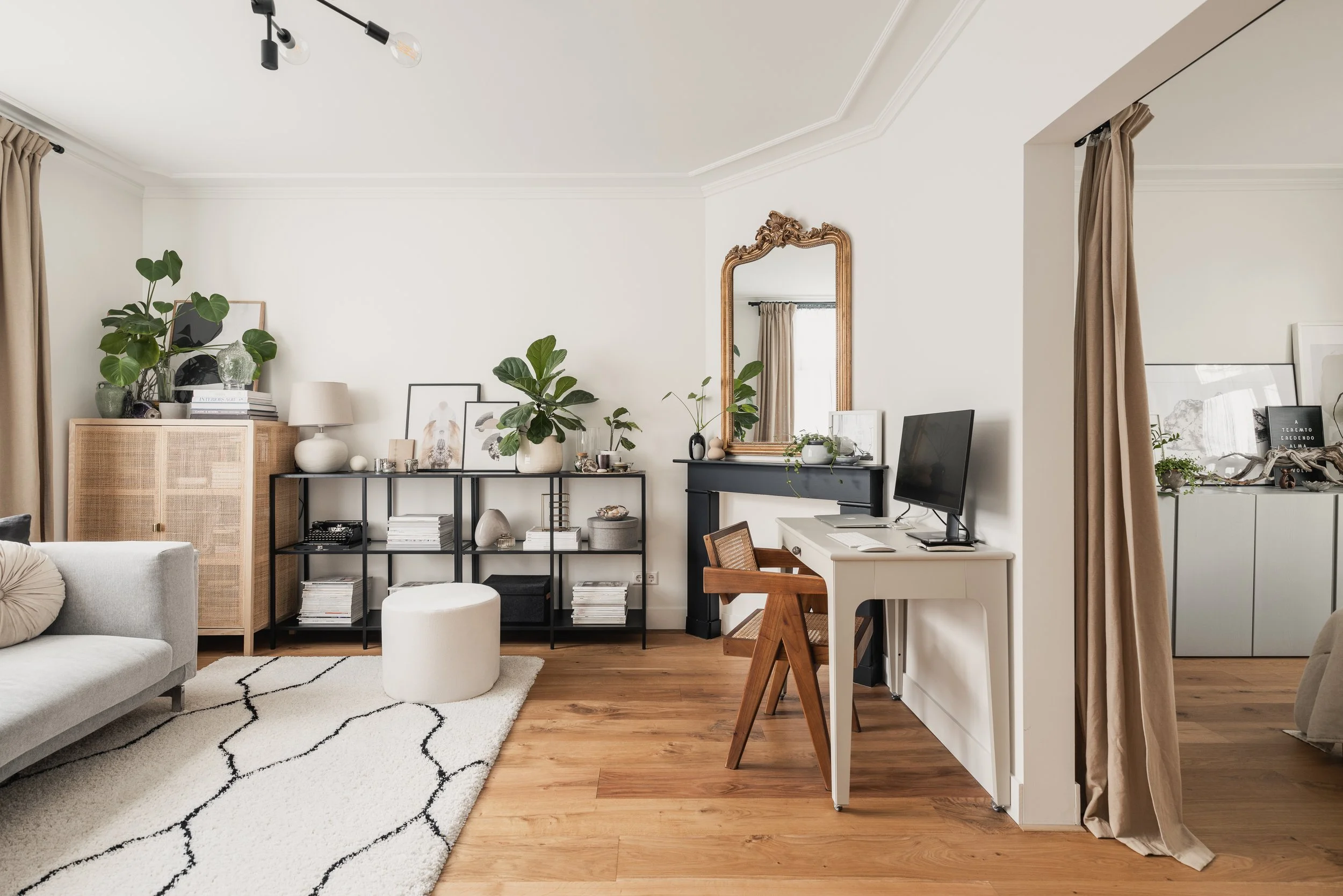Modern living room with white walls, tan curtains, wooden floors, black and white furniture, a computer desk, and decorative plants.