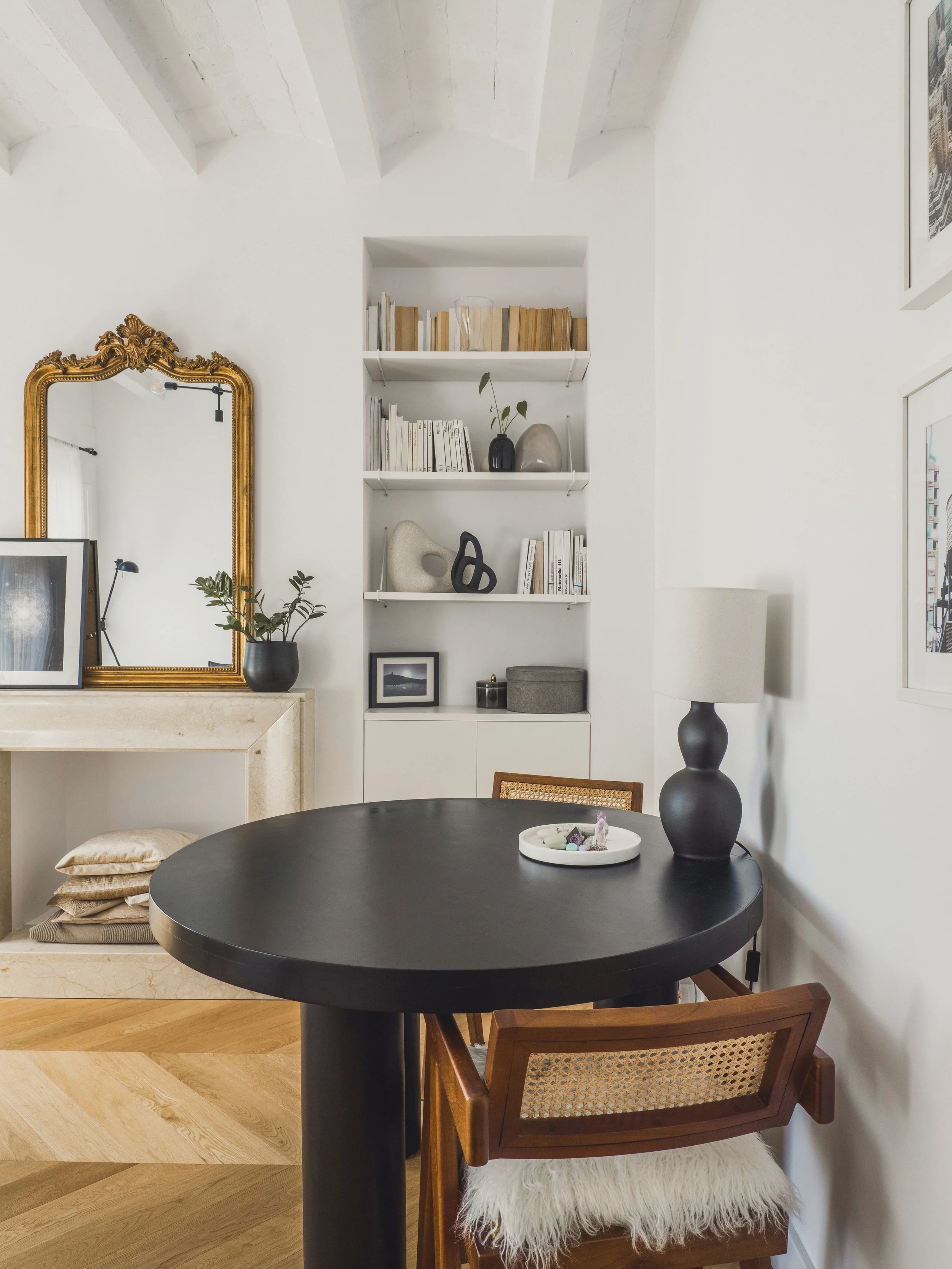 A cozy interior with a round black table, a wooden chair with a white furry cushion, a black table lamp, a shelf with decorative objects and books, a large mirror with an ornate gold frame, and framed artwork on white walls.