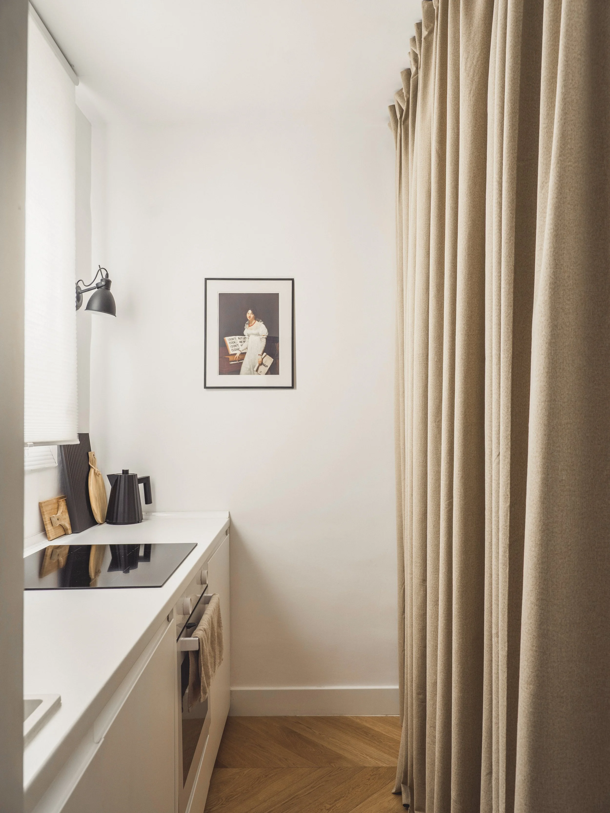 A small kitchen nook with white cabinets, a black electric stove, and a white countertop, decorated with framed pictures, black vases, and wooden accessories. Beige curtains cover the right side, and a framed portrait hangs on the white wall.