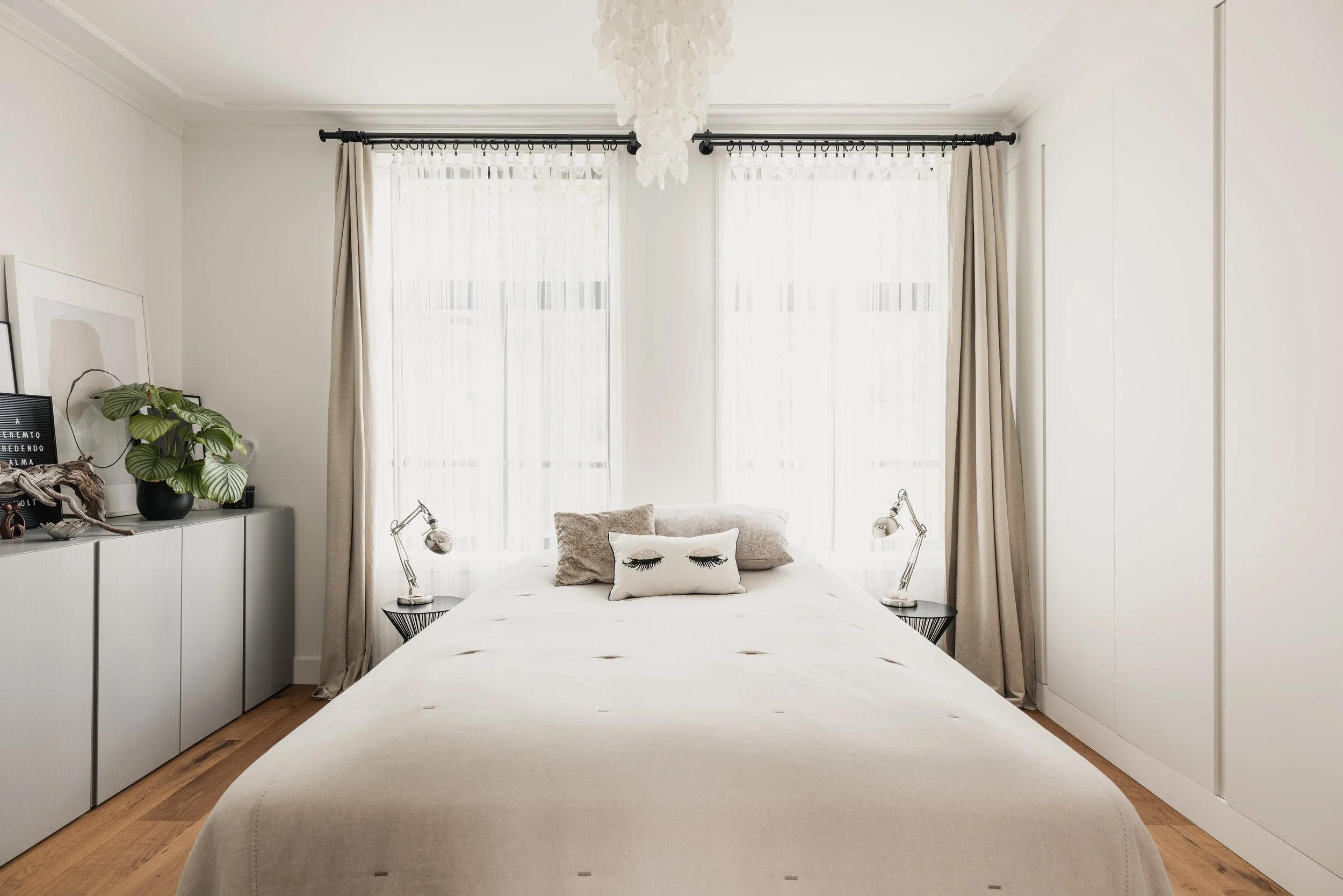 A bright, minimalistic bedroom with a white bed, decorative pillows, two bedside tables with silver lamps, large windows with sheer curtains, a cabinet with plants and pictures, and beige curtains.