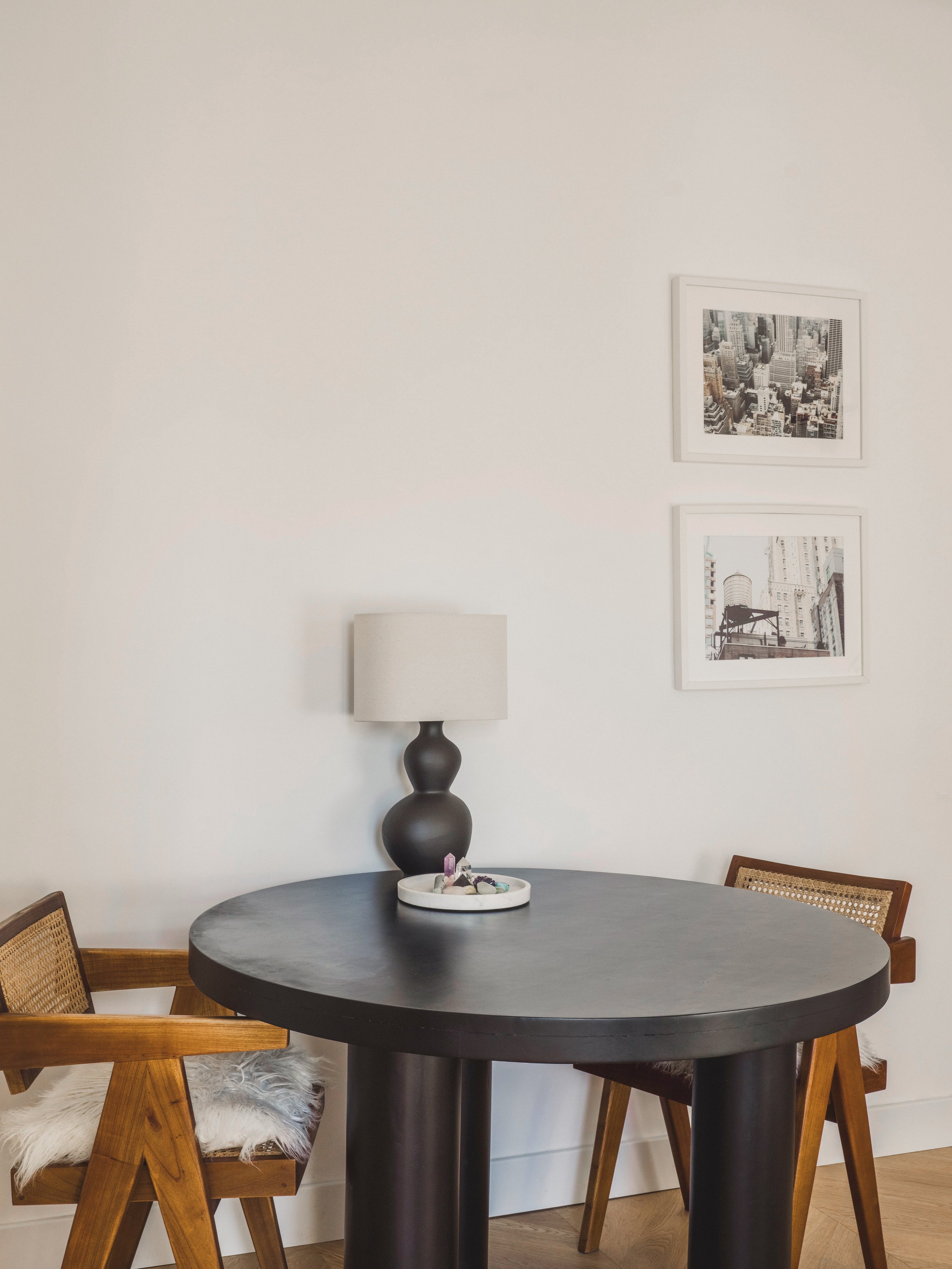 A round black dining table with three wooden chairs, one of which has a white fluffy cushion. On the table, there is a white tray with various crystals and stones. Behind the table, there is a white wall with two framed black and white cityscape phot