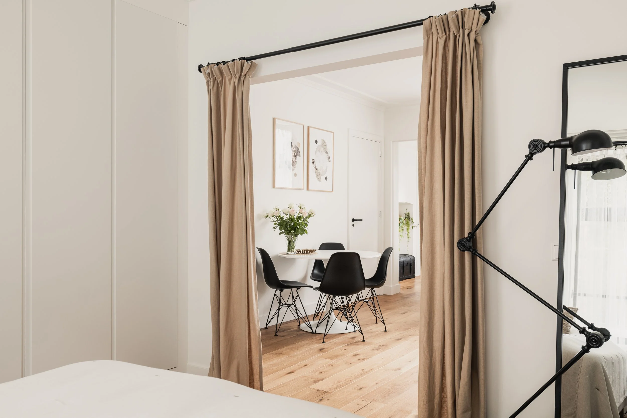 Dining area with a white round table and four black chairs, a vase of flowers on the table, wall art, and a beige curtain framing a view from a bedroom.