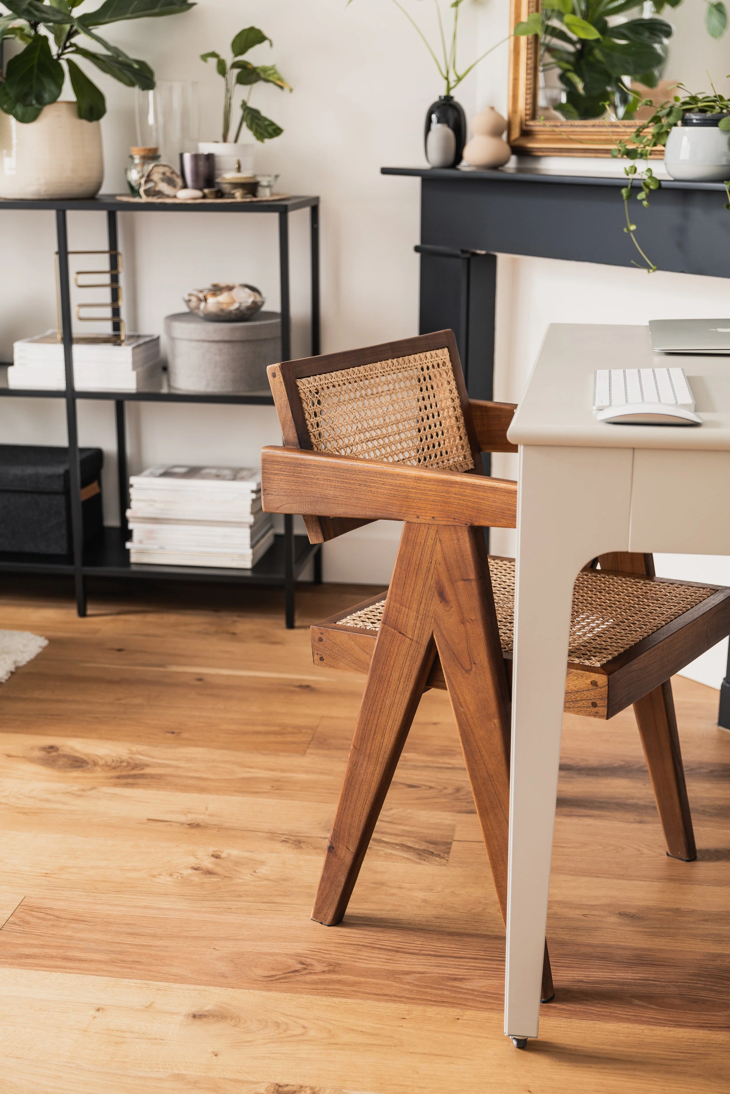 A modern home office with a white desk, wooden chair with woven cane seat and backrest, black shelving unit with plants and decorative objects, and a fireplace mantel decorated with vases and a mirror, on a wooden floor.