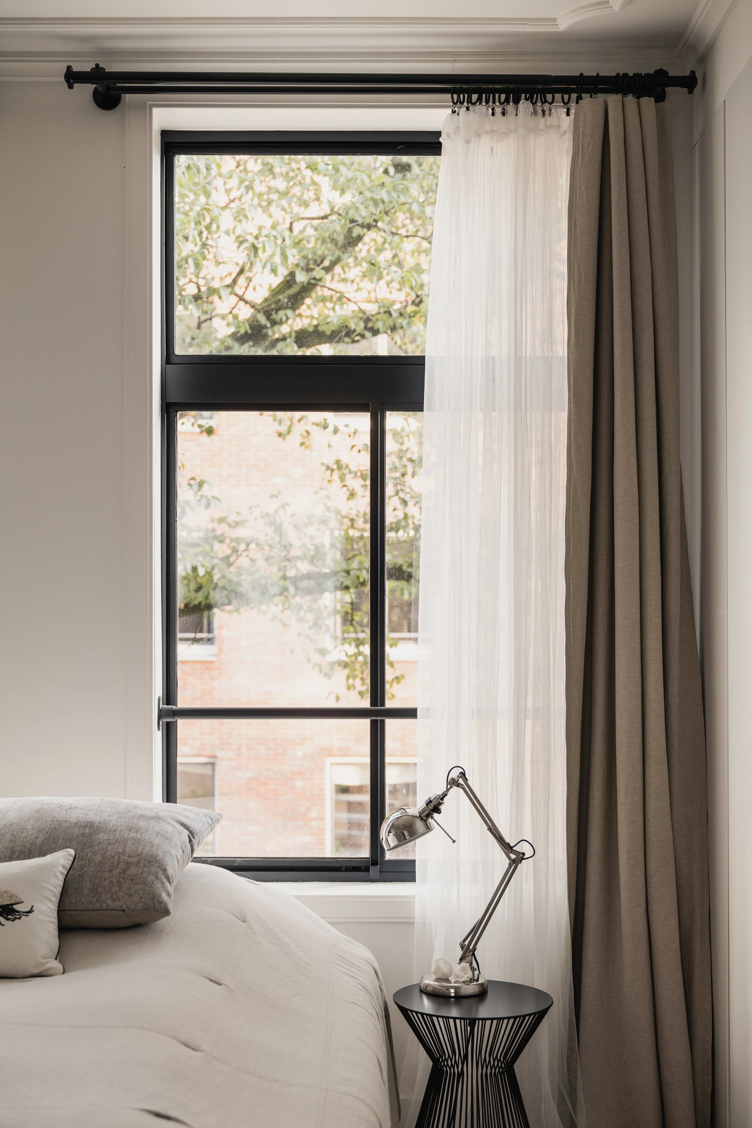 Close-up of a bedroom with a portion of a bed, a black side table, a metallic adjustable desk lamp, and a window with sheer white curtains and beige drapes, showing trees and brick buildings outside.