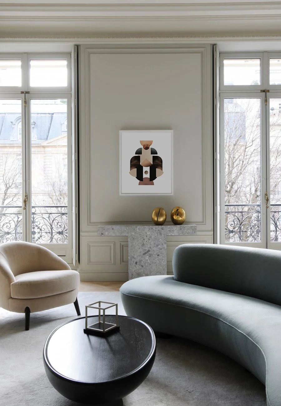 Modern living room with beige armchair, light gray curved sofa, black coffee table with a geometric sculpture, and artwork with abstract faces on the wall. Large windows with wrought iron balcony railing and two gold decorative objects on a marble co