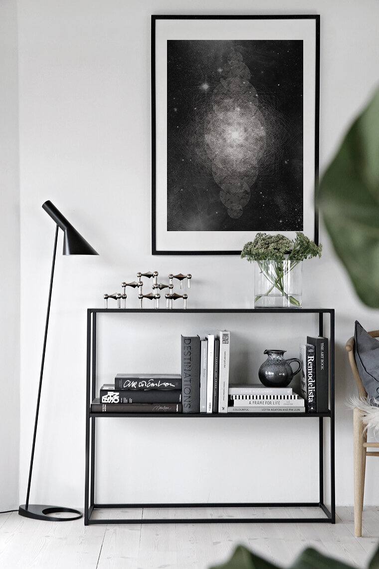 Modern interior with a black metal bookshelf holding books, a black pitcher, and a vase with green flowers, a black angled floor lamp, and a large framed cosmic or galaxy artwork on the white wall.