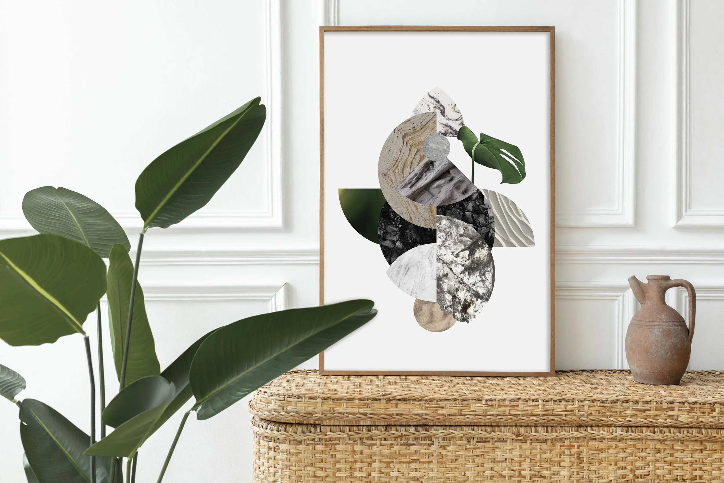 Decorative art print with abstract shapes and natural textures leaning against white paneled wall, next to a terracotta pitcher on woven rattan table, with large leafy green plant in the foreground.