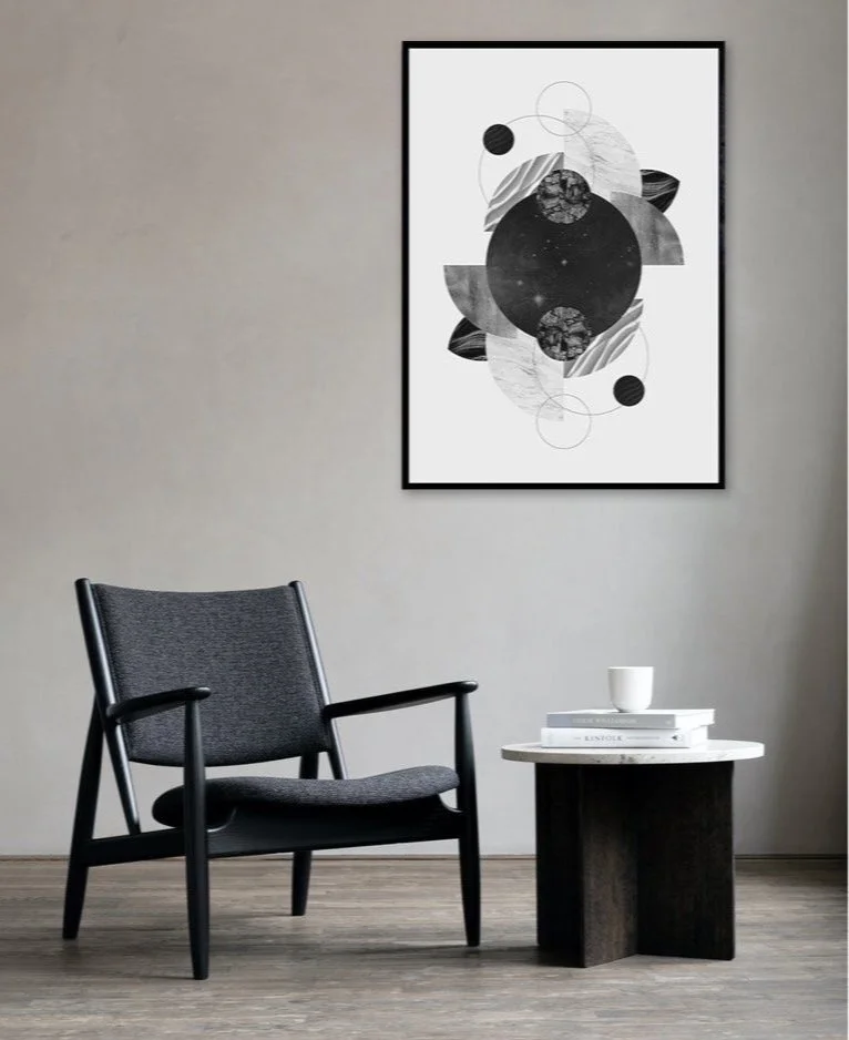 Modern interior with a black armchair, a small round side table with books and a cup, and a geometric black and white abstract art piece on a light gray wall.