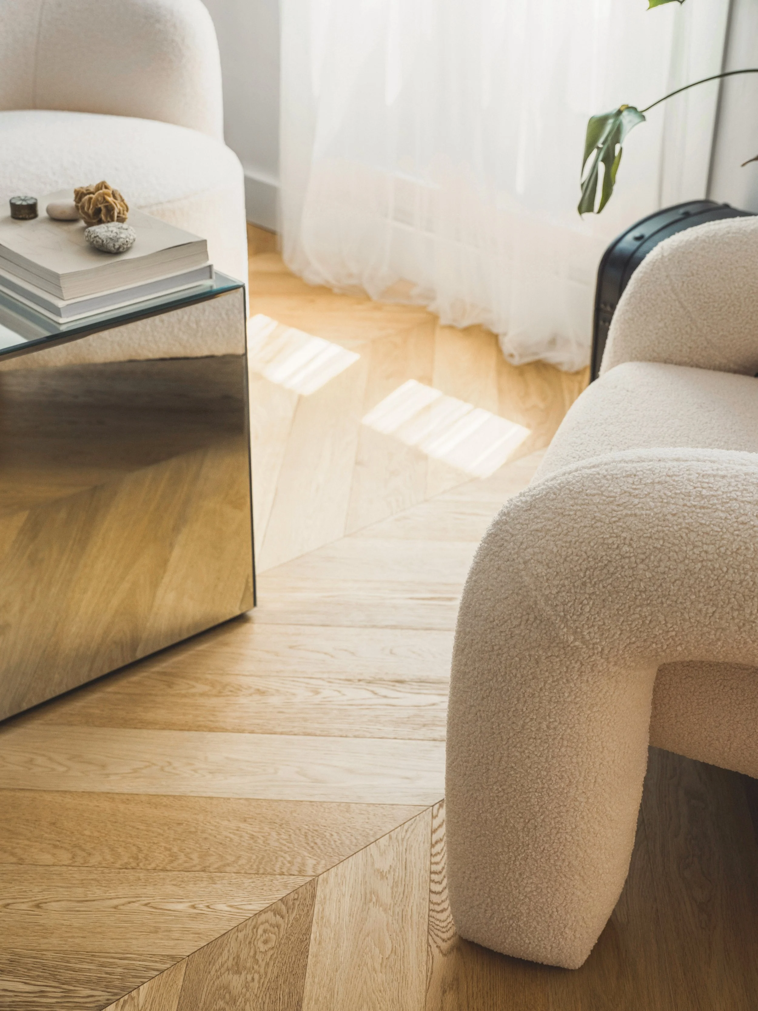 Living room corner with beige textured armchair, wooden floor, and white curtains with sunlight streaming through.