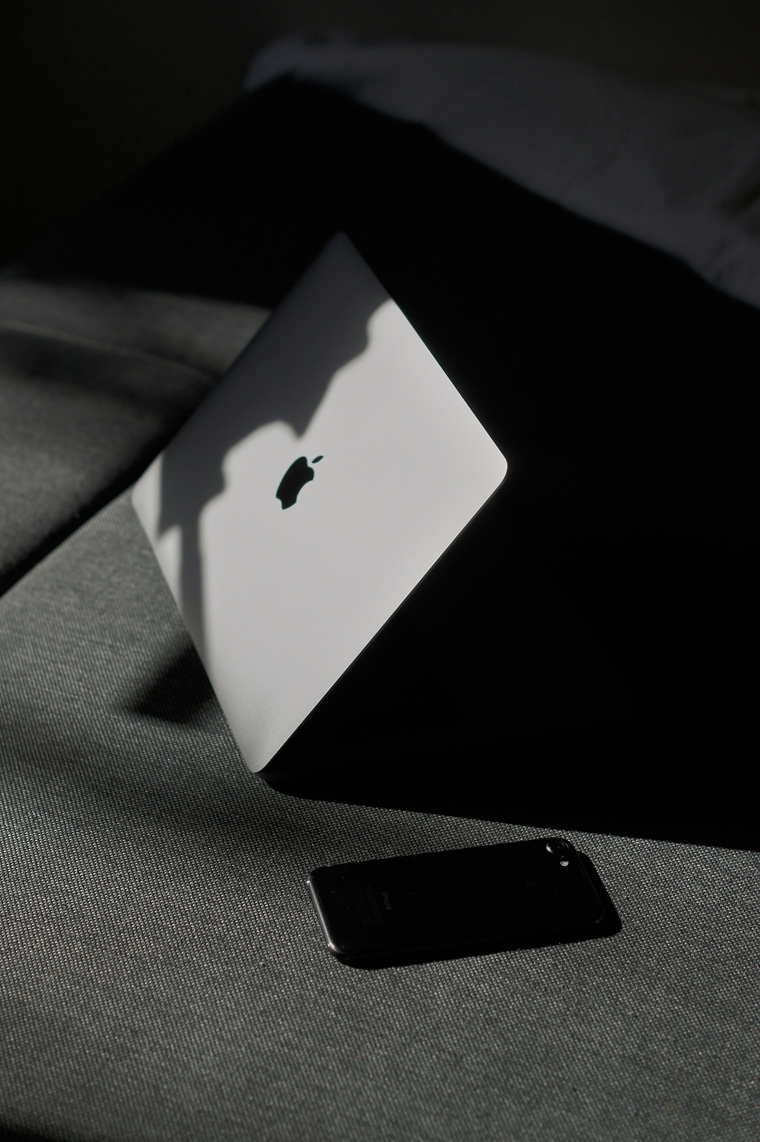 An Apple MacBook laptop and an iPhone placed on a textured surface with shadows cast across them.