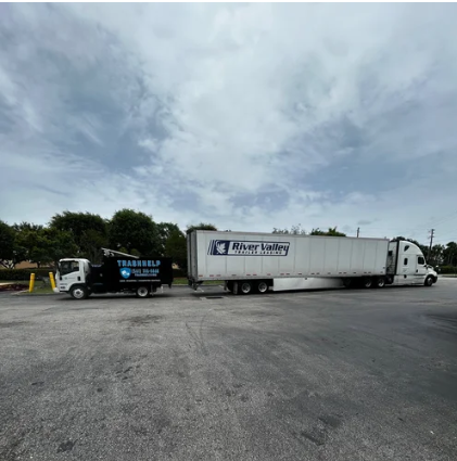 Two semi-trucks parked side by side in a large parking lot with trees and cloudy sky in the background.