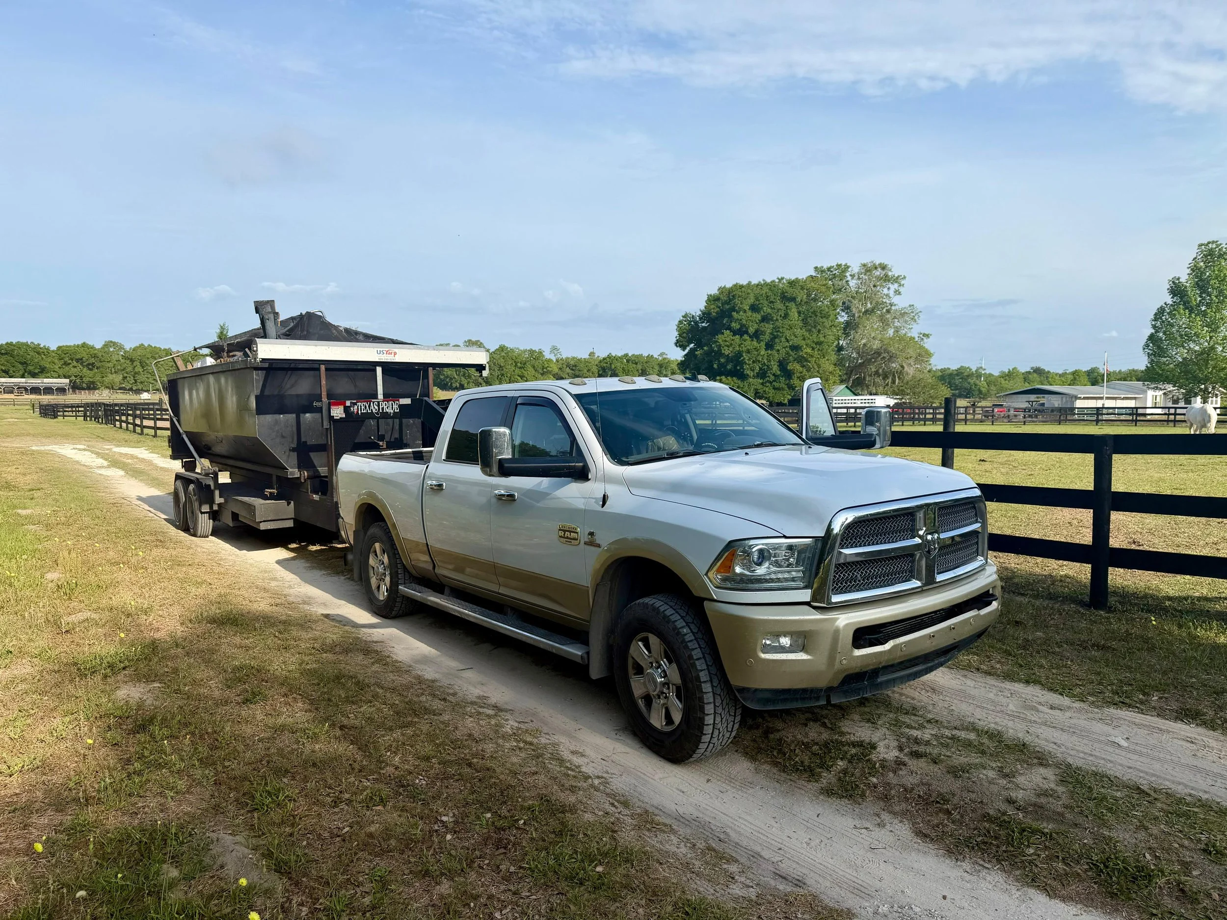 A silver pickup truck towing a black trailer with a large piece of equipment on top, parked on a dirt path in a rural area with fencing, trees, and a cloudy sky.