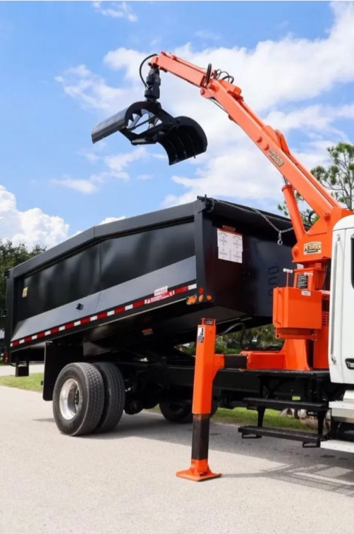 A black dump truck with an orange crane lifting a black debris container, parked on a street under a partly cloudy sky.