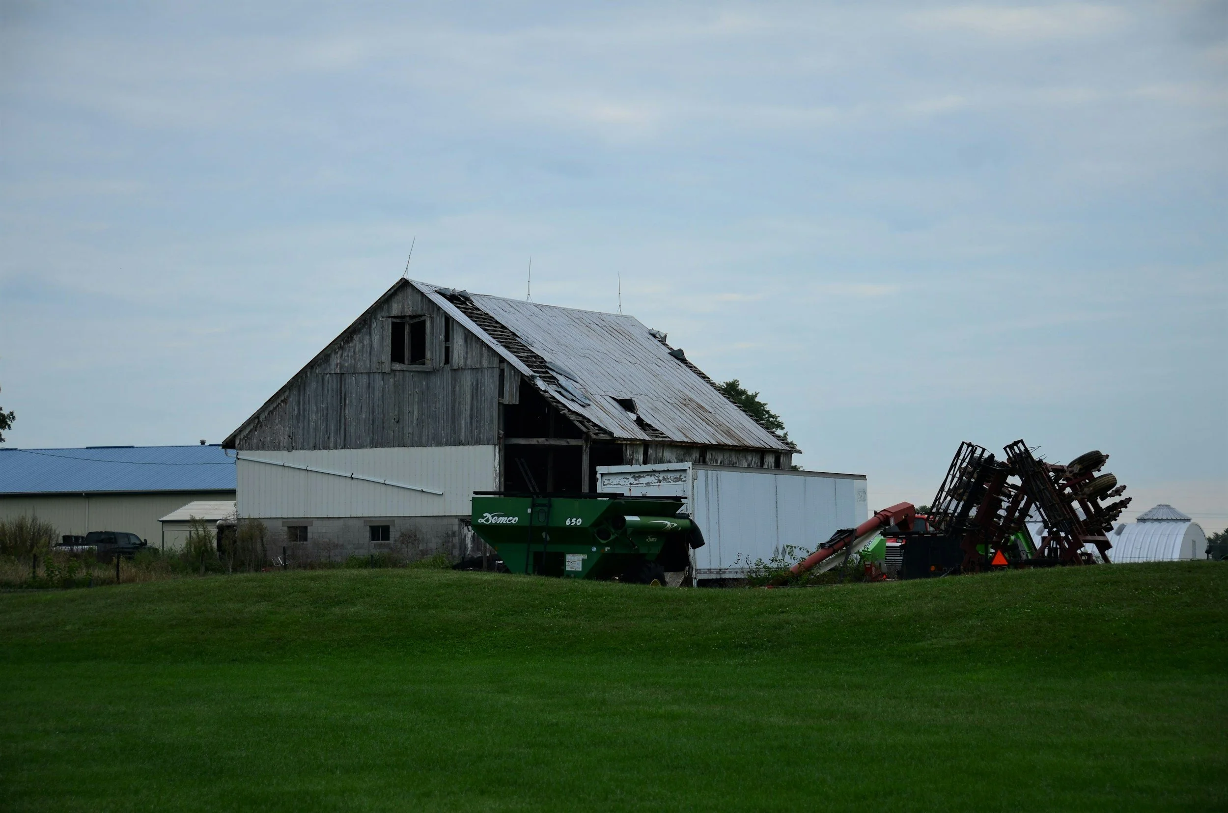 An old wooden barn with a broken roof in a rural farm setting, surrounded by farming equipment and a grassy field under an overcast sky.