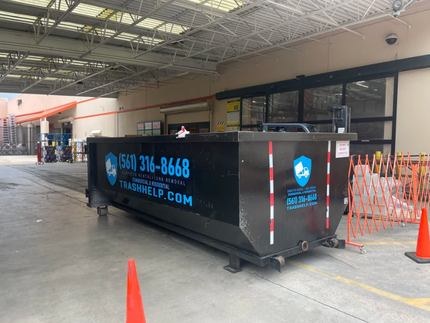 A large black dumpster labeled with blue text and logo for trash removal services, placed in an industrial or commercial area with safety cones and a metal barricade nearby.
