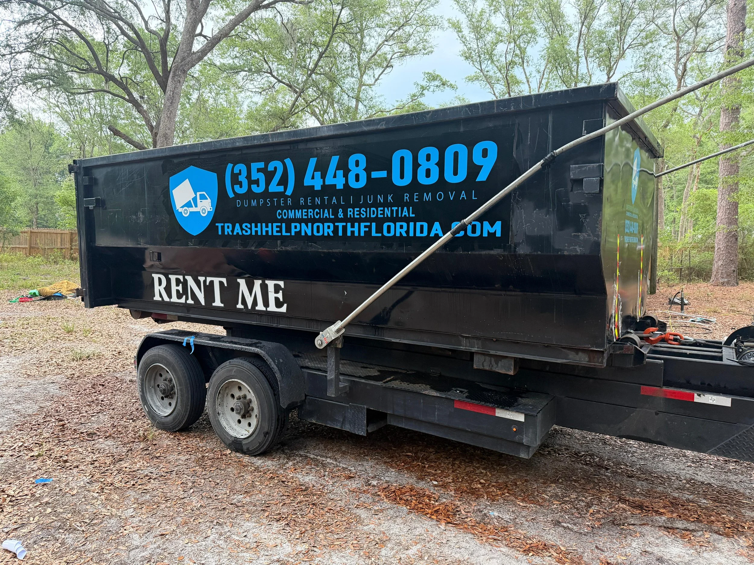 Black trailer with blue lettering advertising dumpster rental and junk removal services, parked outdoors among trees.