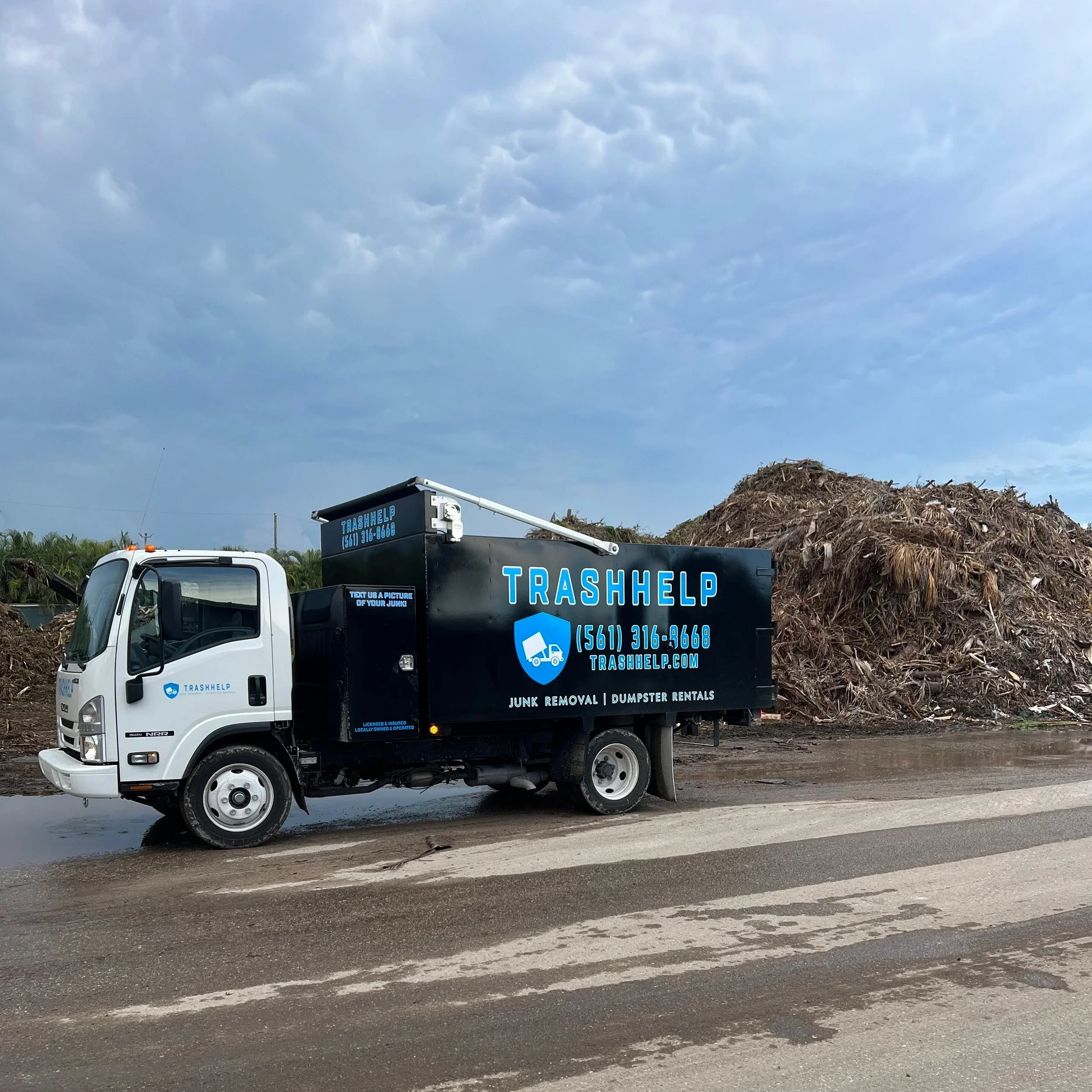 A white truck with a black and blue trash collection container on a road, with a large pile of yard debris and dirt behind it, under a cloudy sky.